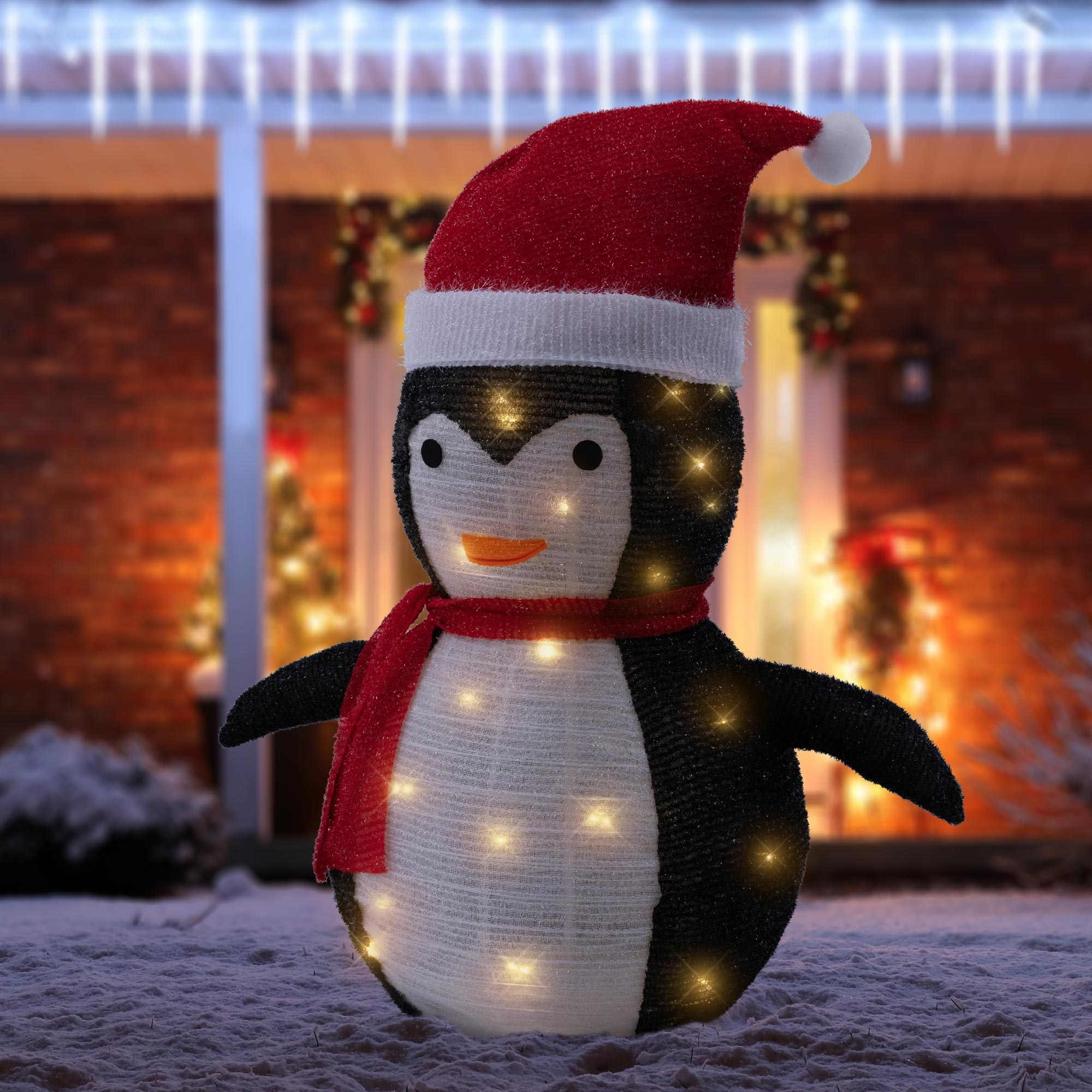 Eine Pinguin-Weihnachtsdekoration mit rotem Hut und Schal, beleuchtet mit Lichtern. Er steht im Schnee.