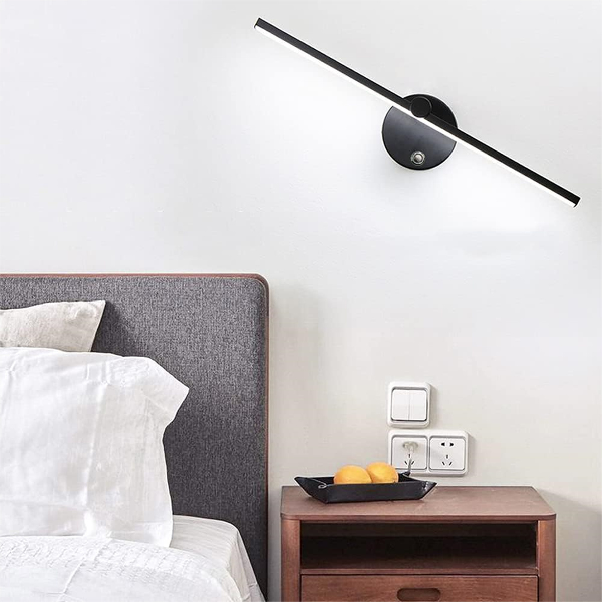 Schlafzimmerwand mit moderner Uhr, Bett, Nachttisch und Steckdosen. Orangen in einer Schale auf dem Nachttisch.