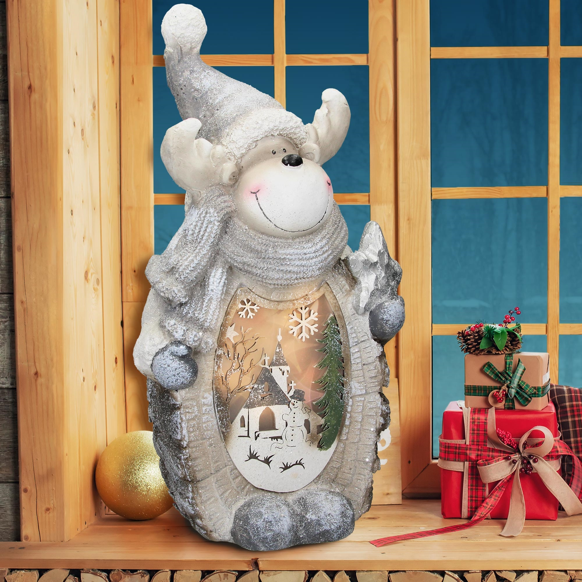 Eine Rentierstatue mit einer Weihnachtsszene im Inneren. Geschenke, eine goldene Kugel und ein Holzfenster im Hintergrund.