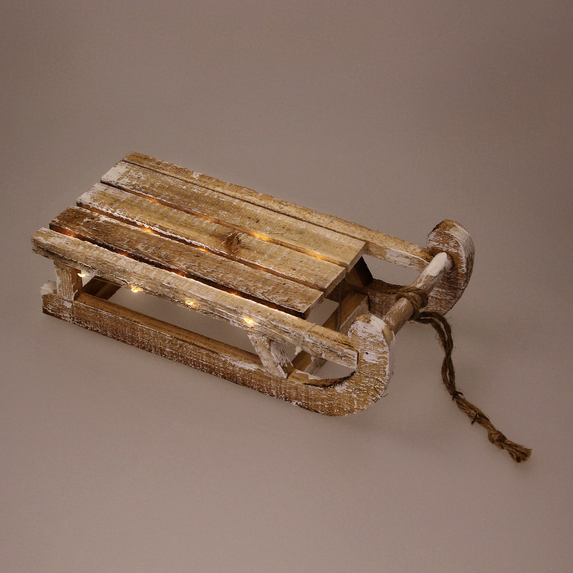 Ein rustikaler Holzschlitten mit weißer Farbe und Schnur auf hellbraunem Hintergrund, der mit kleinen Lichtern beleuchtet wird.