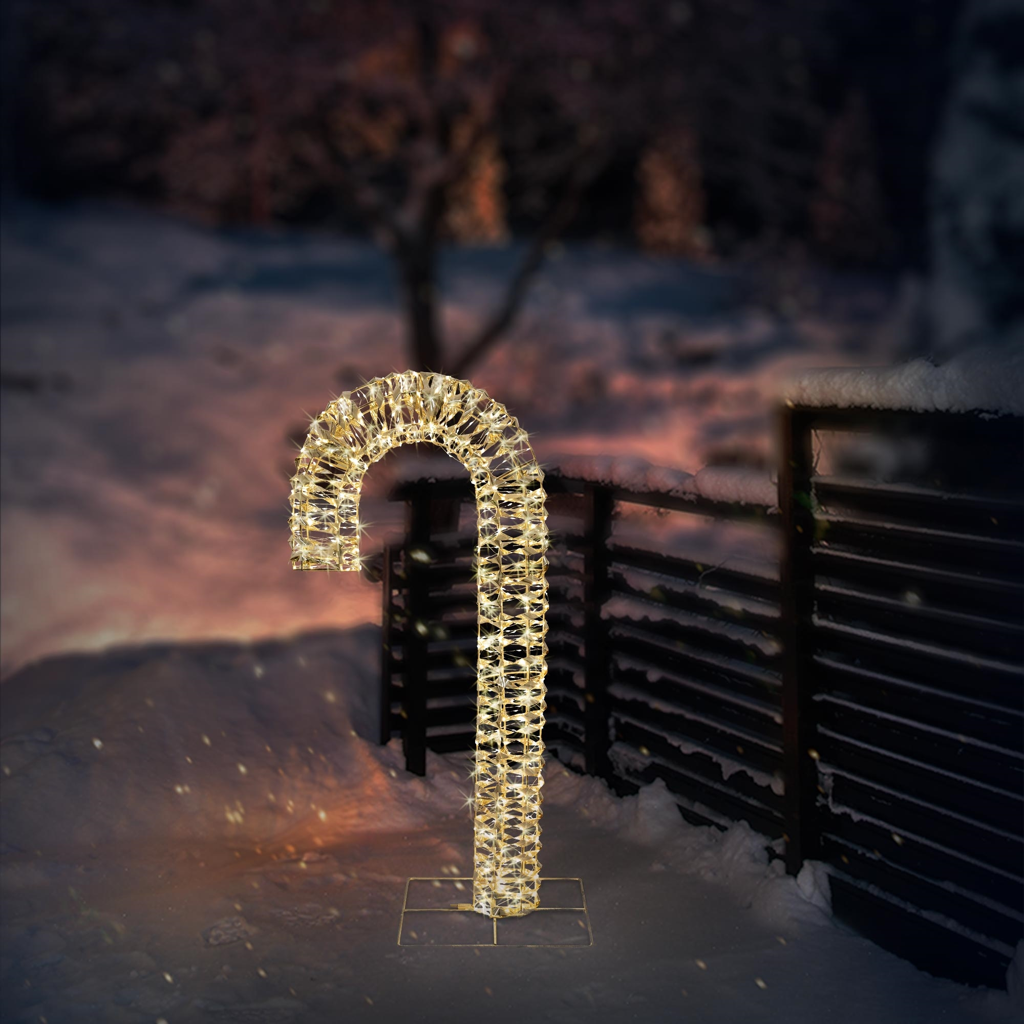 Eine beleuchtete Zuckerstangen-Dekoration steht im Schnee. Im Hintergrund sind ein Zaun und verschwommene Bäume. Die Stange besteht aus Lichtern.
