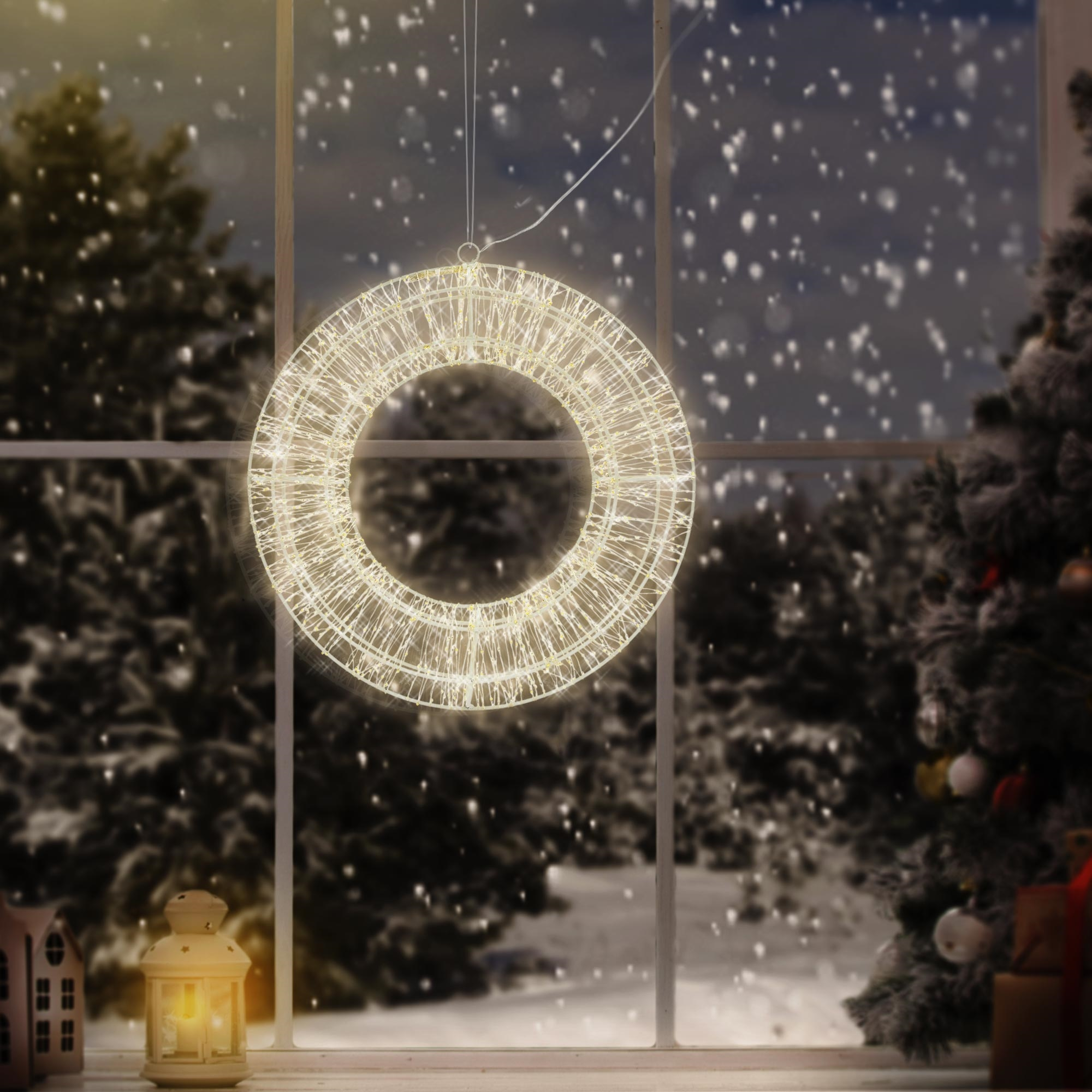Ein beleuchteter Weihnachtskranz hängt vor einem schneebedeckten Fenster. Eine beleuchtete Laterne und ein Baum stehen auf der Fensterbank.