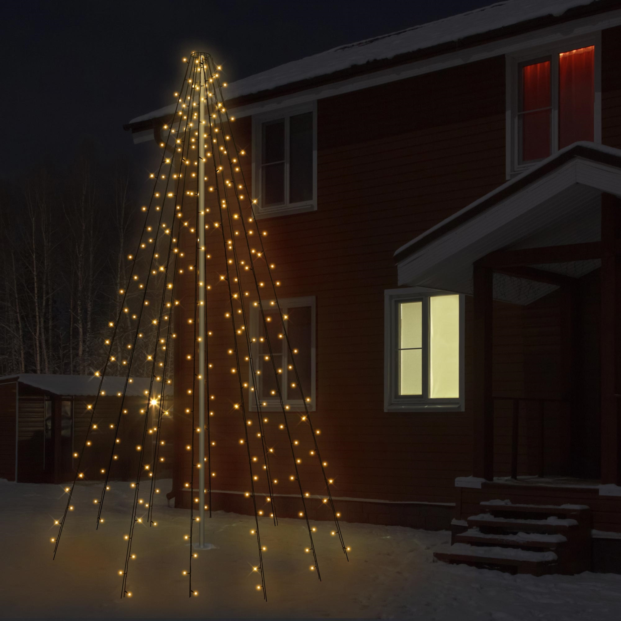 Eine beleuchtete, weihnachtsbaumförmige Anzeige steht nachts vor einem Haus, umgeben von Schnee. Fenster des Hauses sind beleuchtet.