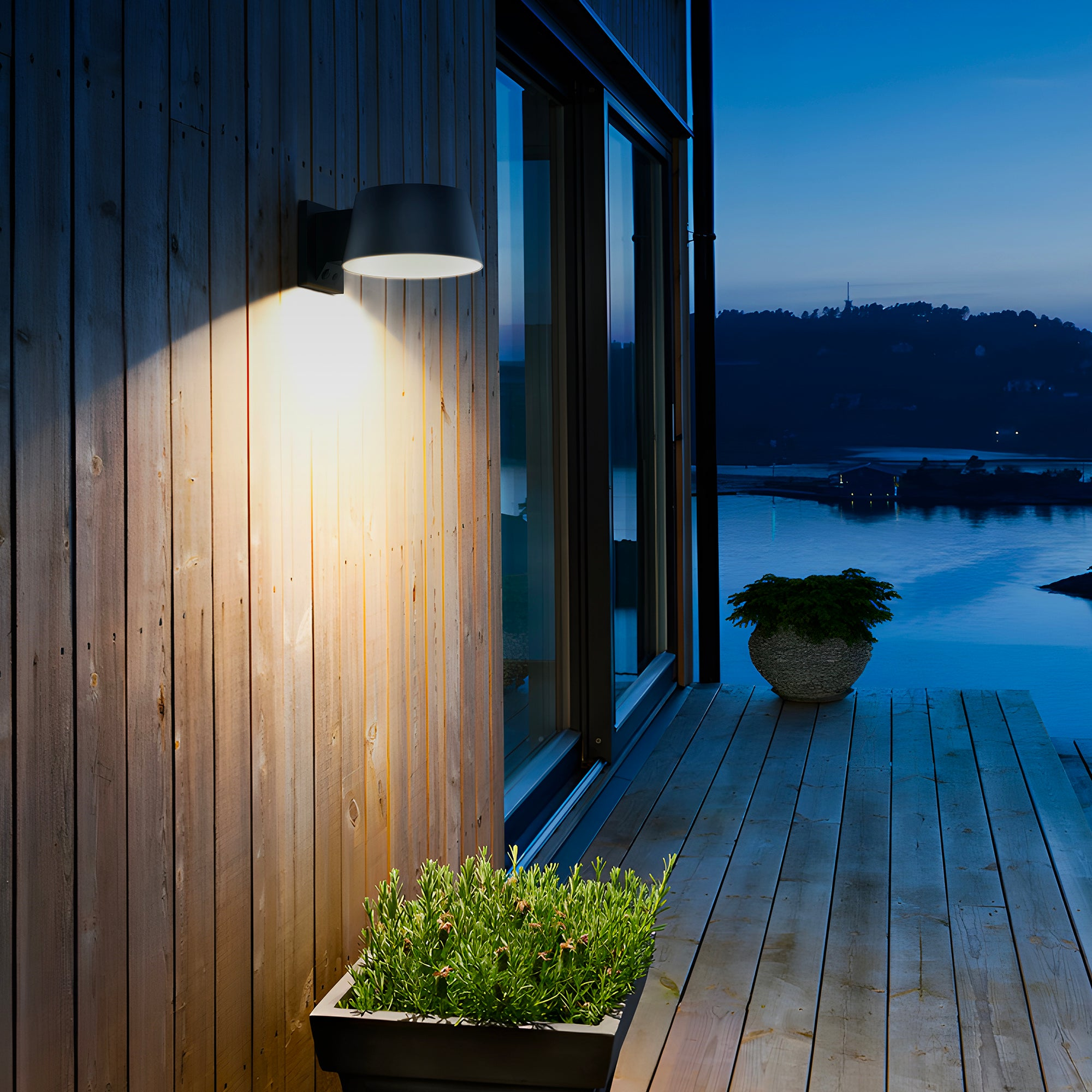 Außenaufnahme in der Dämmerung. Holzwand mit beleuchteter Lampe. Davor ein Blumentopf. Im Hintergrund Wasser und Himmel.