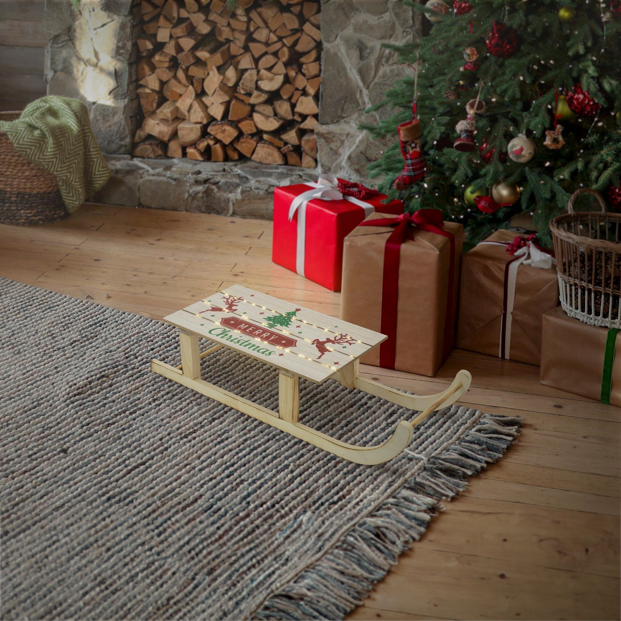 Ein dekorierter Holzschlitten liegt auf einem Teppich. Weihnachtsbaum und verpackte Geschenke im Hintergrund.