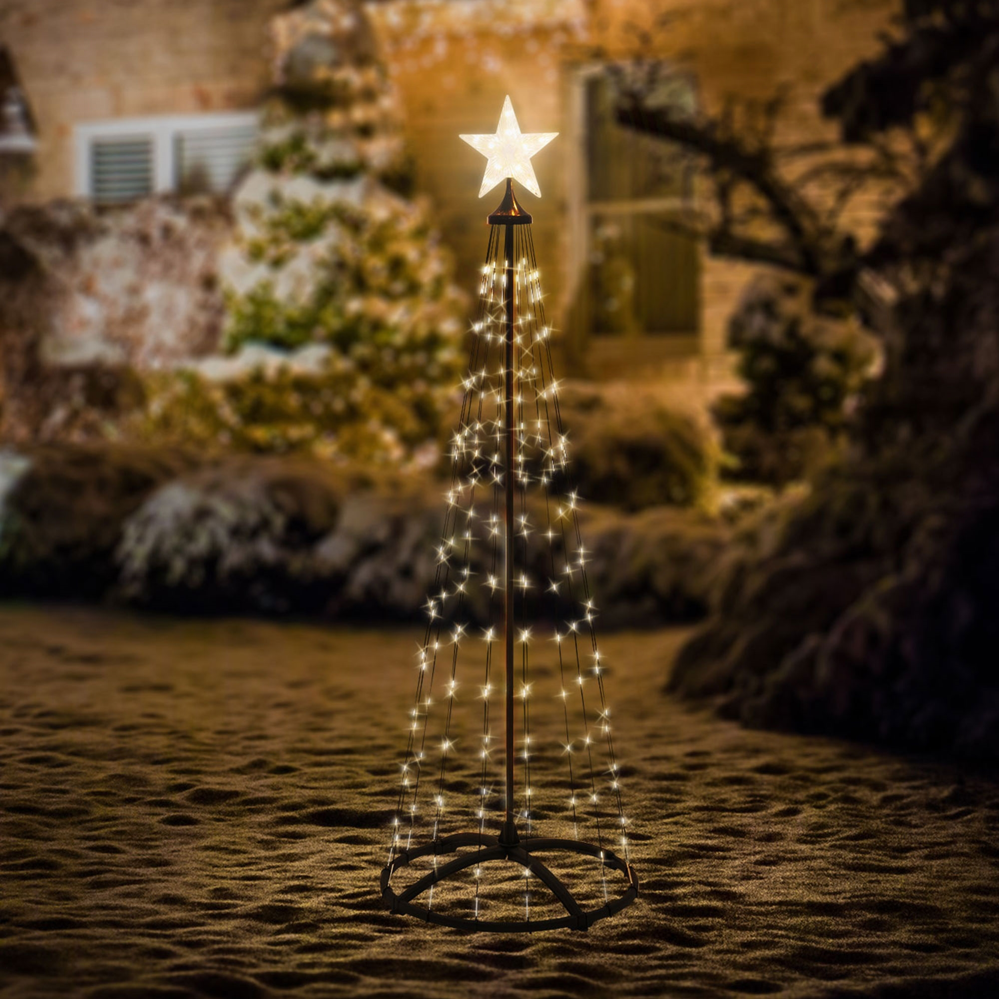 Eine beleuchtete weihnachtsbaumförmige Dekoration mit einem Stern oben steht auf dem Boden. Der Hintergrund zeigt verschwommenes Haus und Büsche.