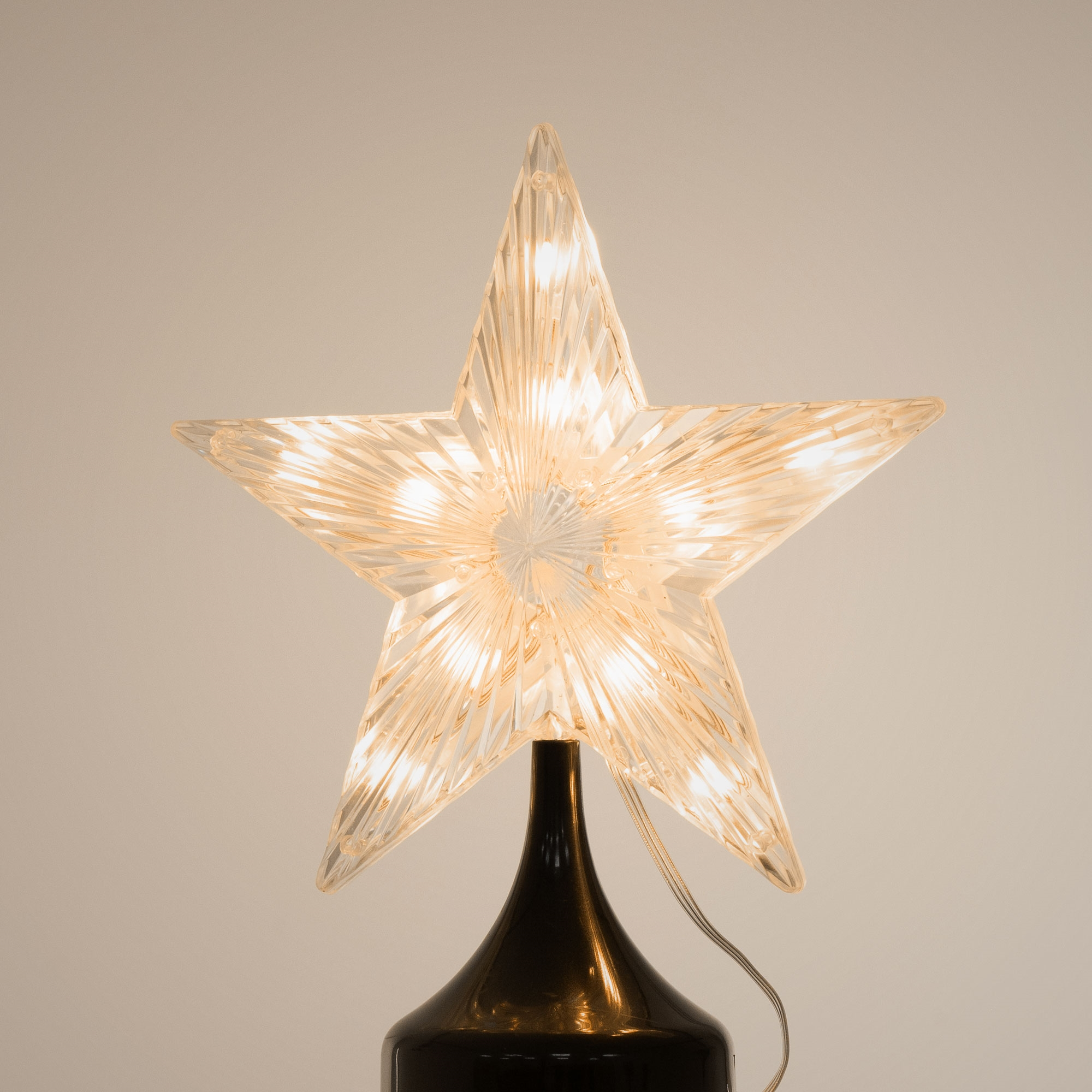 Ein klarer, sternförmiger Weihnachtsbaumaufsatz mit Lichtern. Er steht auf einem schwarzen Sockel vor weißem Hintergrund.
