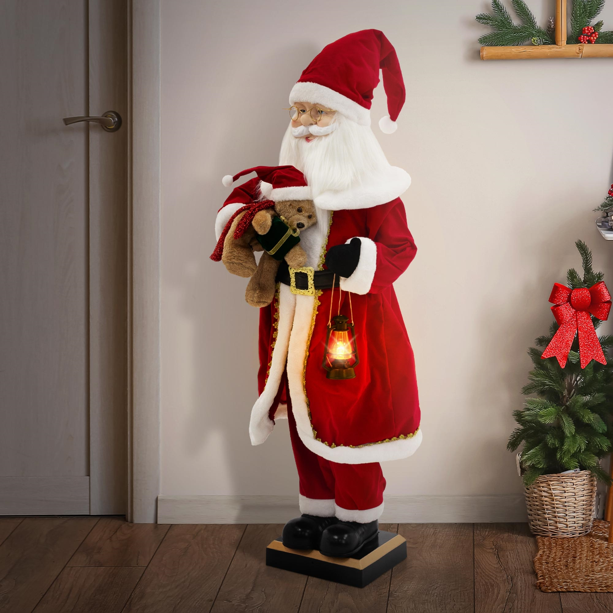 Weihnachtsmann-Statue mit Teddybär und Laterne, trägt einen roten Anzug. Weihnachtsbaum und Tür im Hintergrund.