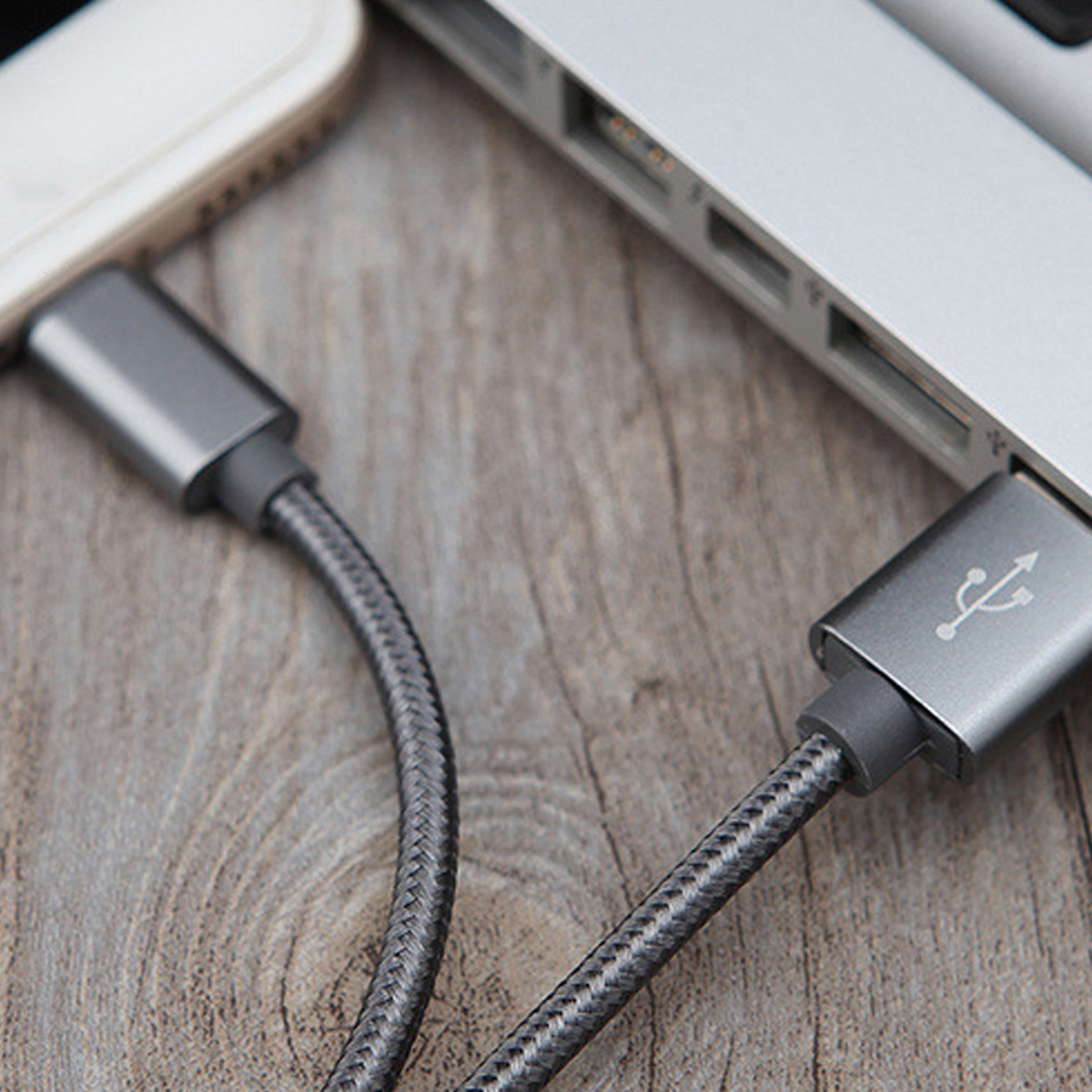 Ein graues USB-Kabel ist mit einem Laptop und einem Telefon verbunden. Der Hintergrund zeigt eine Holzoberfläche.