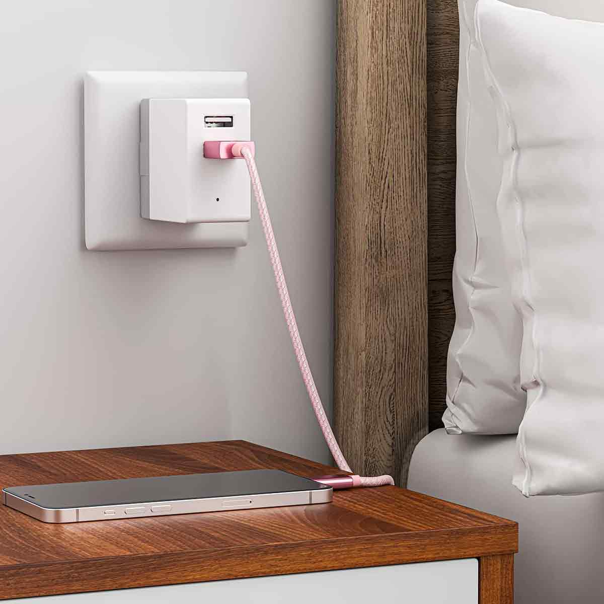 Ein Telefon wird auf einem Nachttisch geladen. Ein rosa Kabel verbindet sich mit einem Wandladegerät. Im Hintergrund ein Bett und ein Nachttisch.