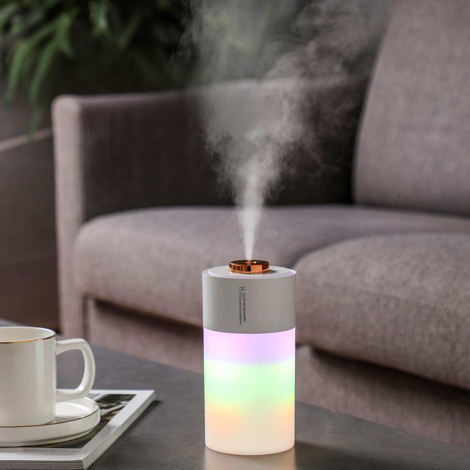 Ein Luftbefeuchter stößt Nebel aus. Er steht auf einem Tisch in der Nähe einer Couch und einer Tasse. Der Luftbefeuchter hat farbwechselnde Lichter.