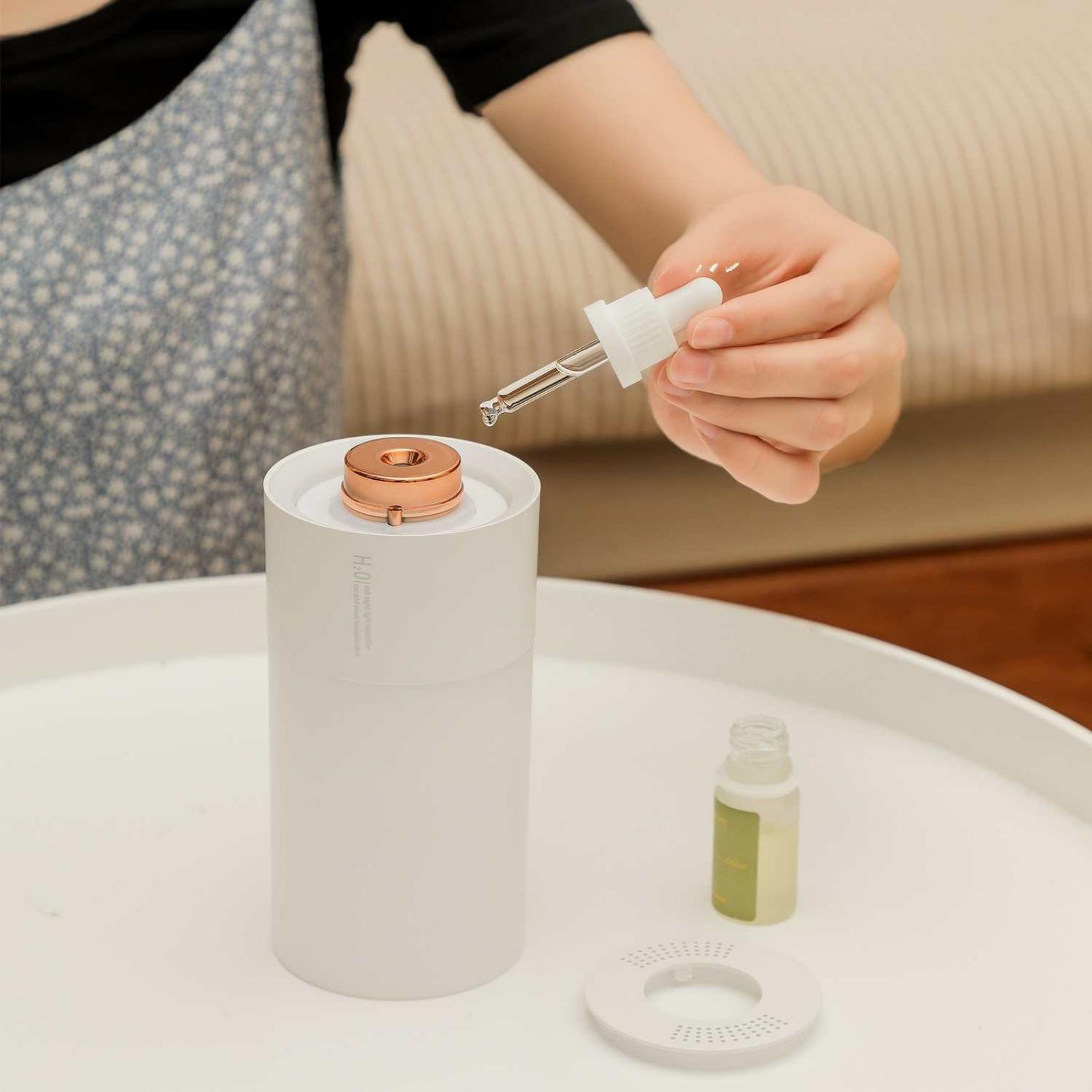 Eine Hand hält eine Pipette und tropft Flüssigkeit in einen weißen Diffusor mit Kupferdeckel. Eine kleine Flasche ist in der Nähe.