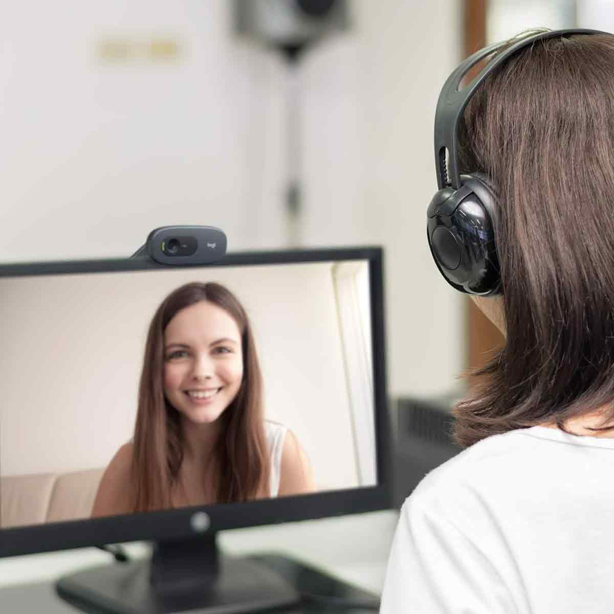 Eine Person mit Kopfhörern und einer Webcam an einem Monitor. Der Bildschirm zeigt eine lächelnde Frau. Die Person ist in einem Videoanruf.