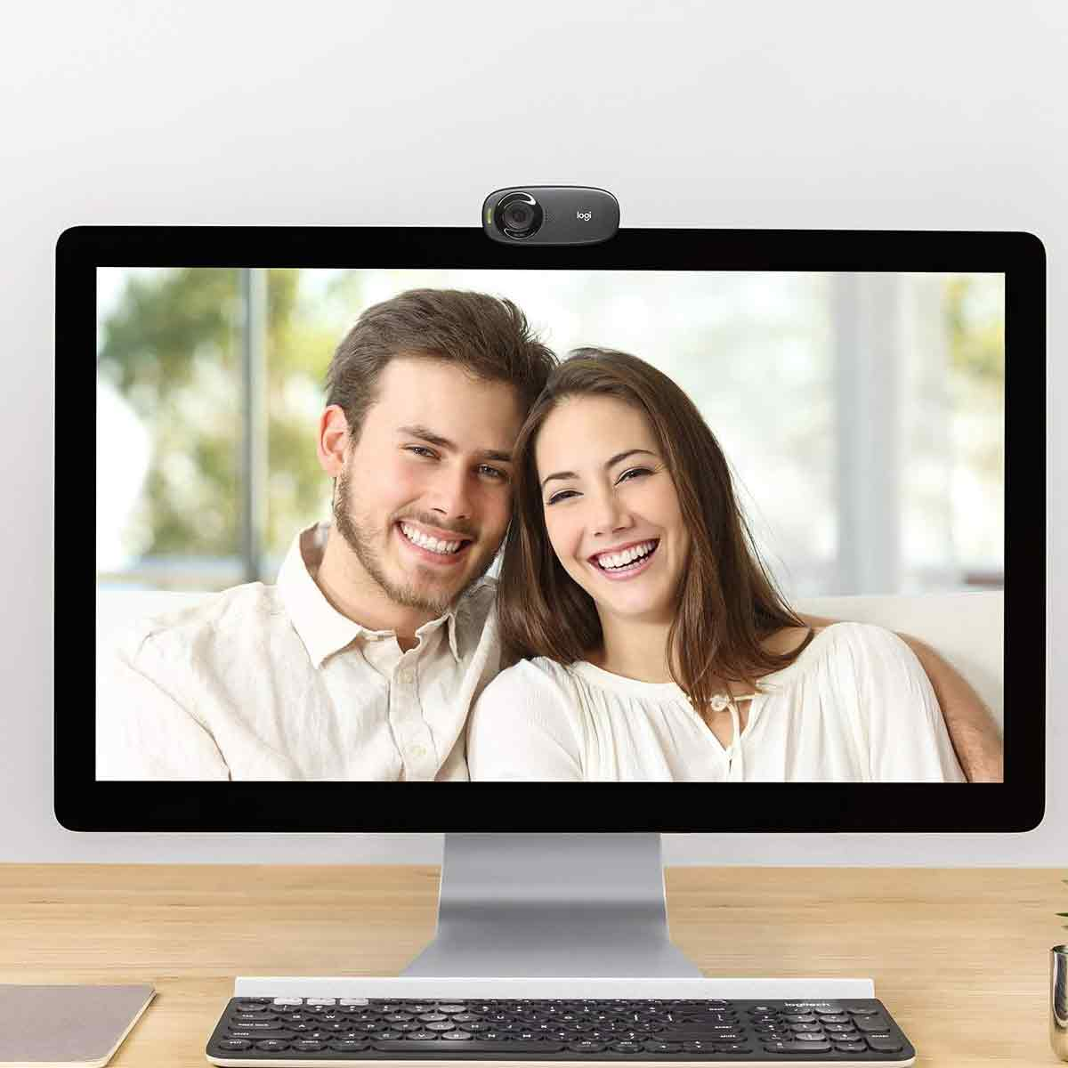 Auf einem Computerbildschirm ist ein lächelndes Paar zu sehen. Oben befindet sich eine Webcam. Unten ist eine Tastatur, der Tisch ist aus Holz.