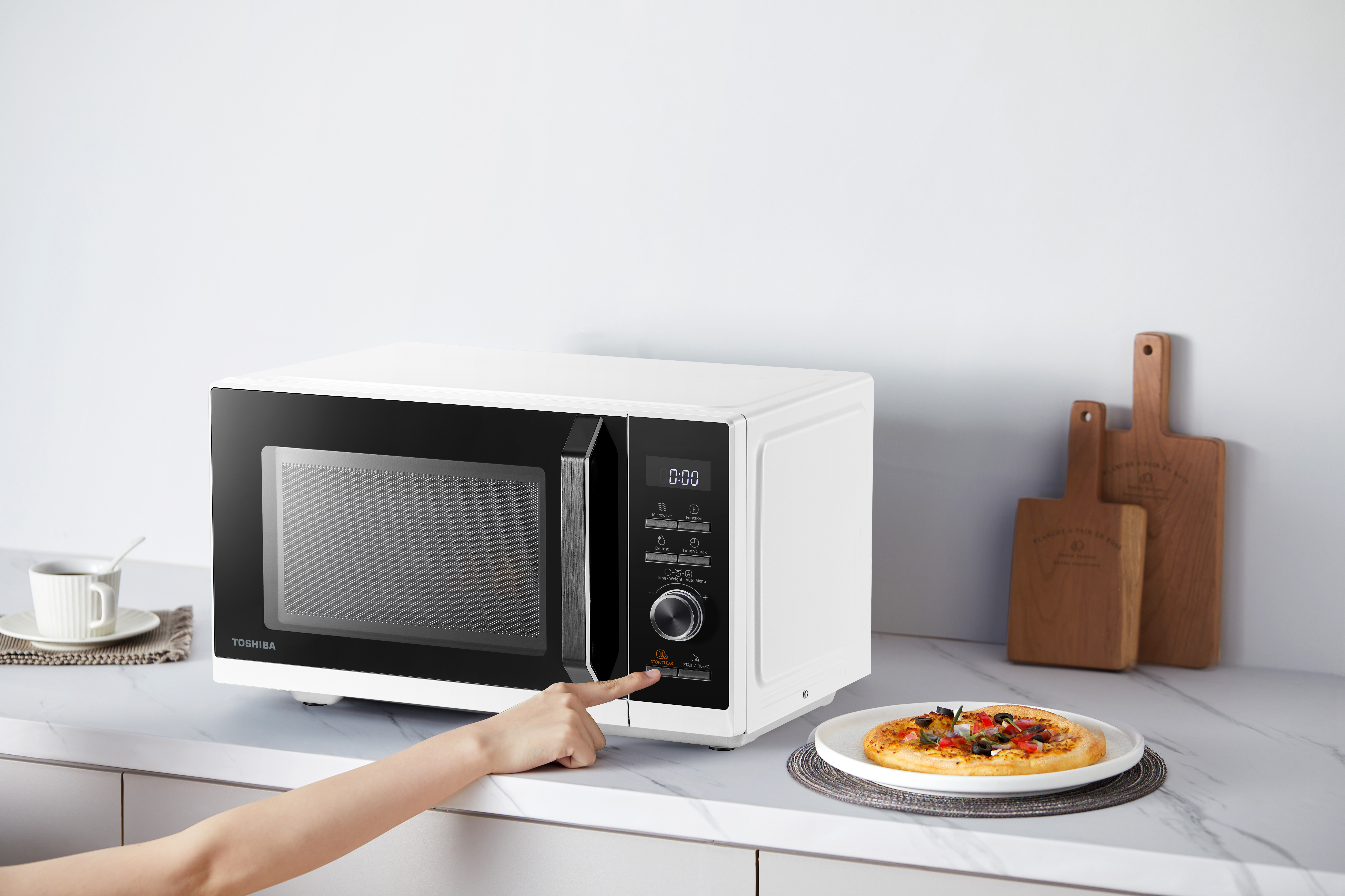 Un forno a microonde bianco Toshiba con una mano che preme un pulsante. Una pizza è sul bancone.