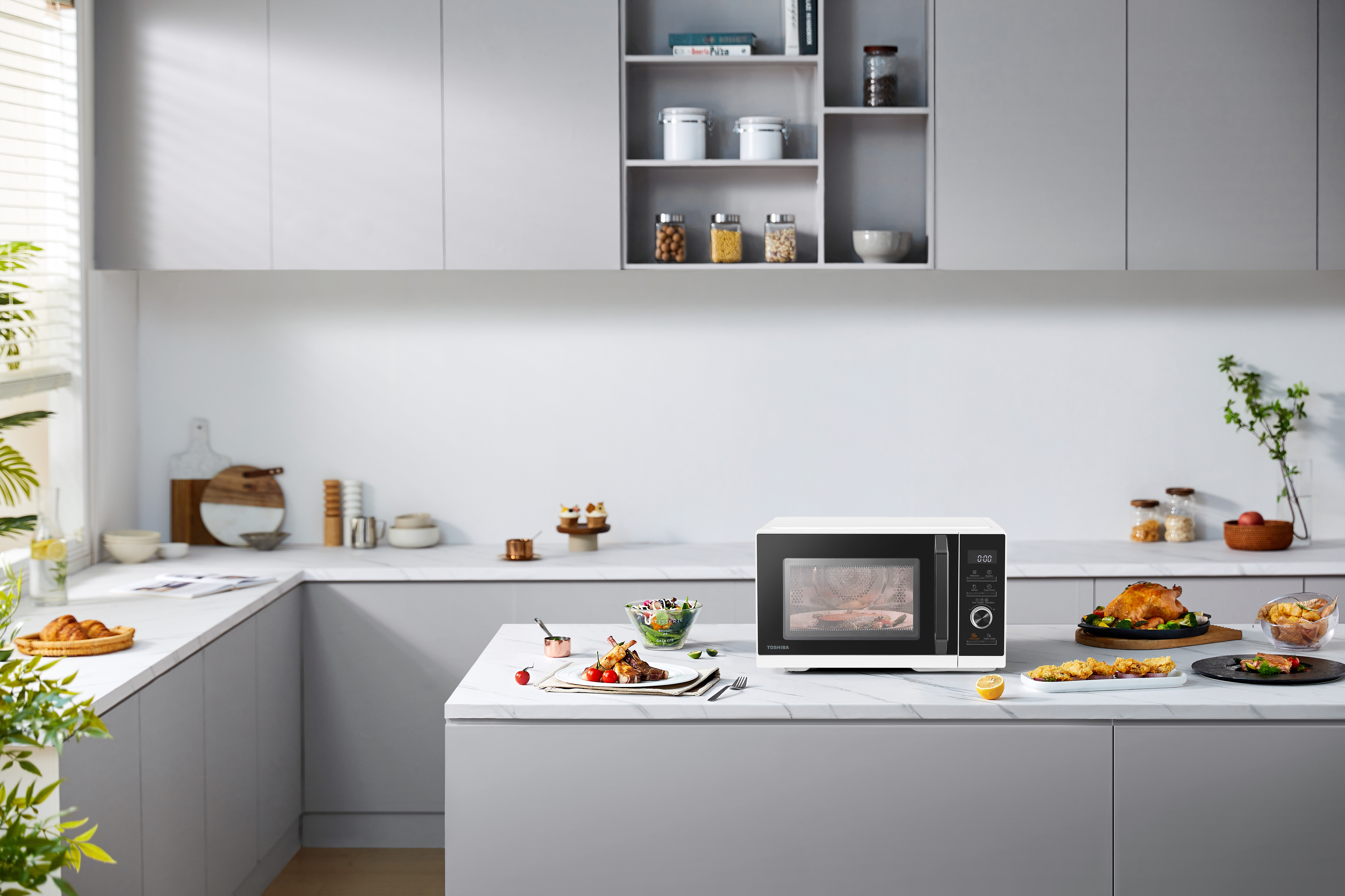 Una scena di cucina con microonde, cibo cotto e vari oggetti da cucina su un piano di lavoro bianco.