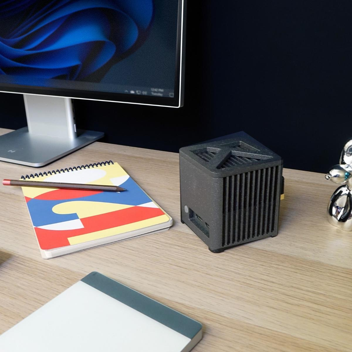 Scena na biurku z komputerem, notatnikiem, ołówkiem i czarnym urządzeniem w kształcie pudełka. Obecna jest również srebrna statua psa z balonów.