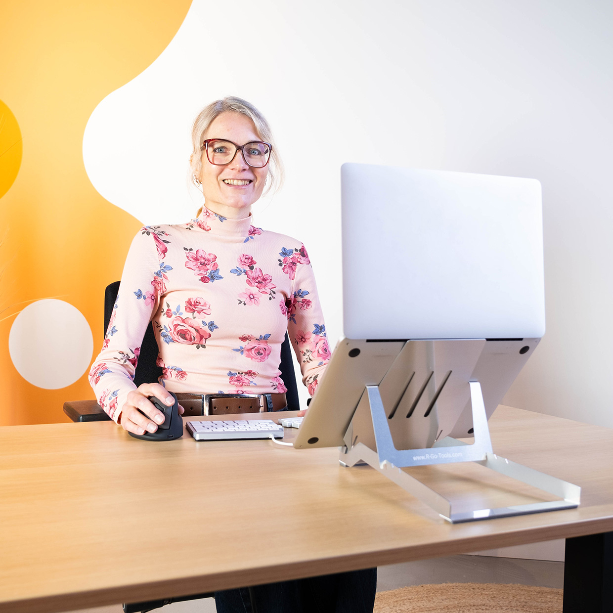 Frau mit Brille lächelt in die Kamera. Sie sitzt an einem Schreibtisch mit Laptop und Maus. Blumenmuster, heller Tisch.