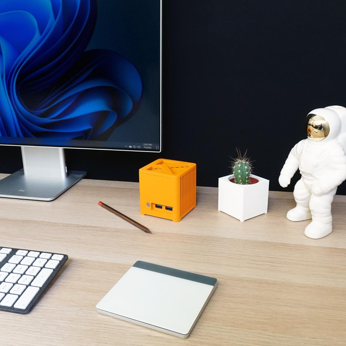 Ustawienie biurka z monitorem pokazującym niebieski abstrakcyjny wzór, pomarańczową kostką, ołówkiem i białą figurką astronauty.