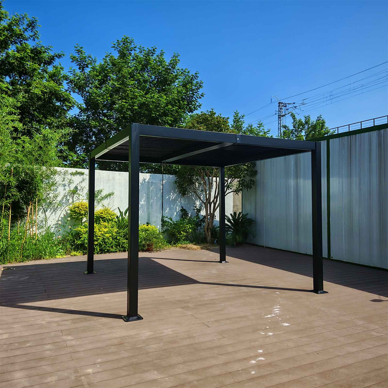 Eine schwarze Pergola steht auf einem Holzdeck, umgeben von Grün unter einem klaren blauen Himmel. Im Hintergrund ist eine weiße Wand.