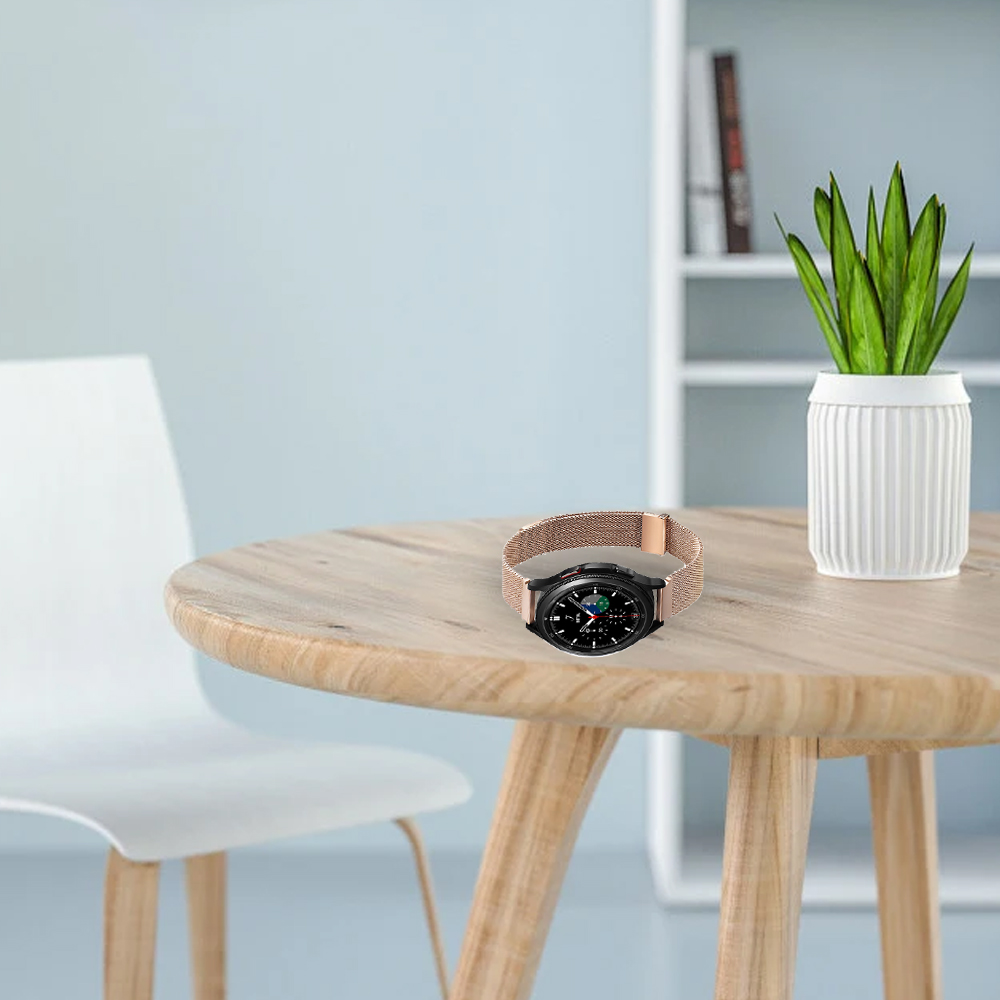 Eine Uhr mit beigem Armband liegt auf einem Holztisch. Links steht ein weißer Stuhl. Rechts eine Pflanze in einem weißen Topf.
