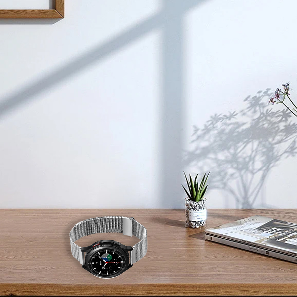 Eine Smartwatch mit silbernem Armband liegt auf einem Holztisch. Eine kleine Pflanze und eine Zeitschrift liegen ebenfalls auf dem Tisch. Der Hintergrund ist weiß.