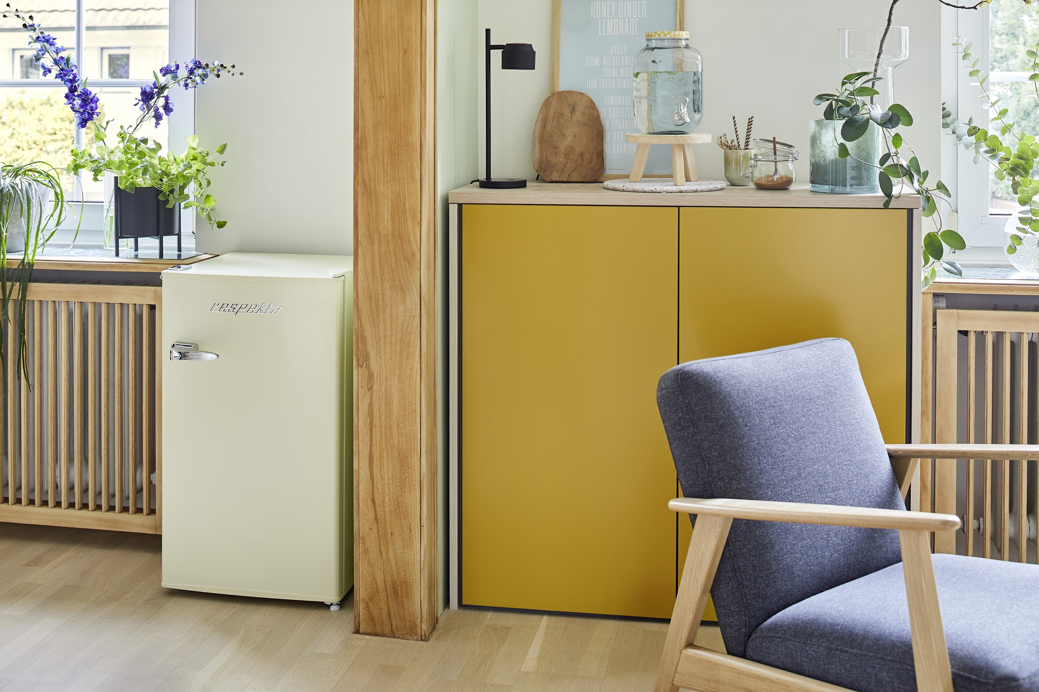 Zimmer mit Kühlschrank, gelbem Schrank und blauem Stuhl. Pflanzen und Dekorationen auf dem Schrank. Holzboden und Heizkörper.