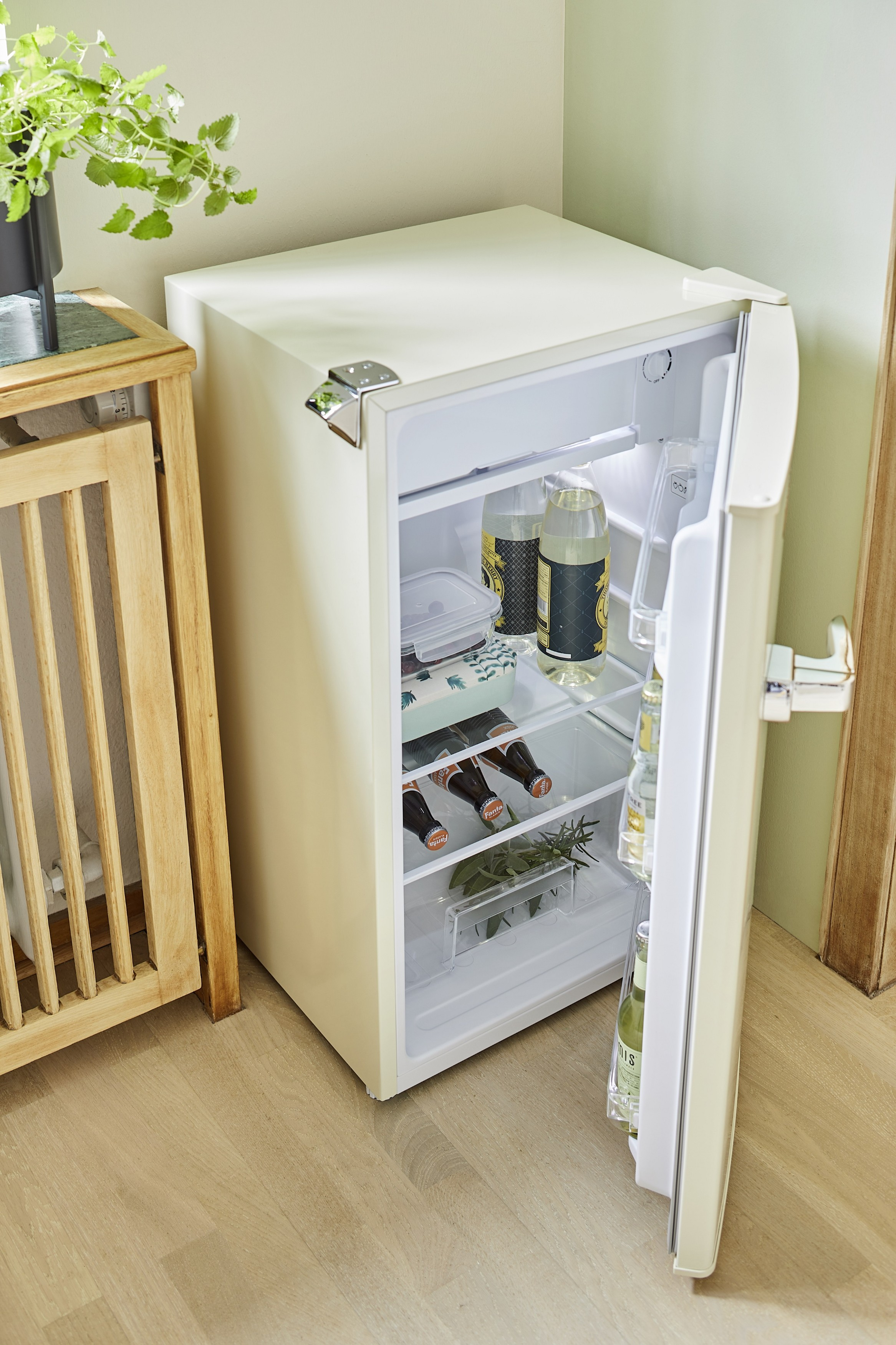 Offener Kühlschrank mit Getränken, neben Holztisch mit Pflanze und Holztor.