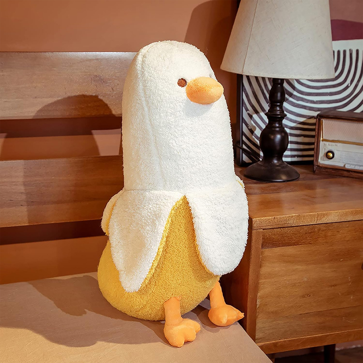 Ein Plüschtier in Form einer Bananenente. Es hat einen weißen Bananenschalenkopf, einen gelben Körper, einen orangefarbenen Schnabel und Füße. Holzmöbel im Hintergrund.