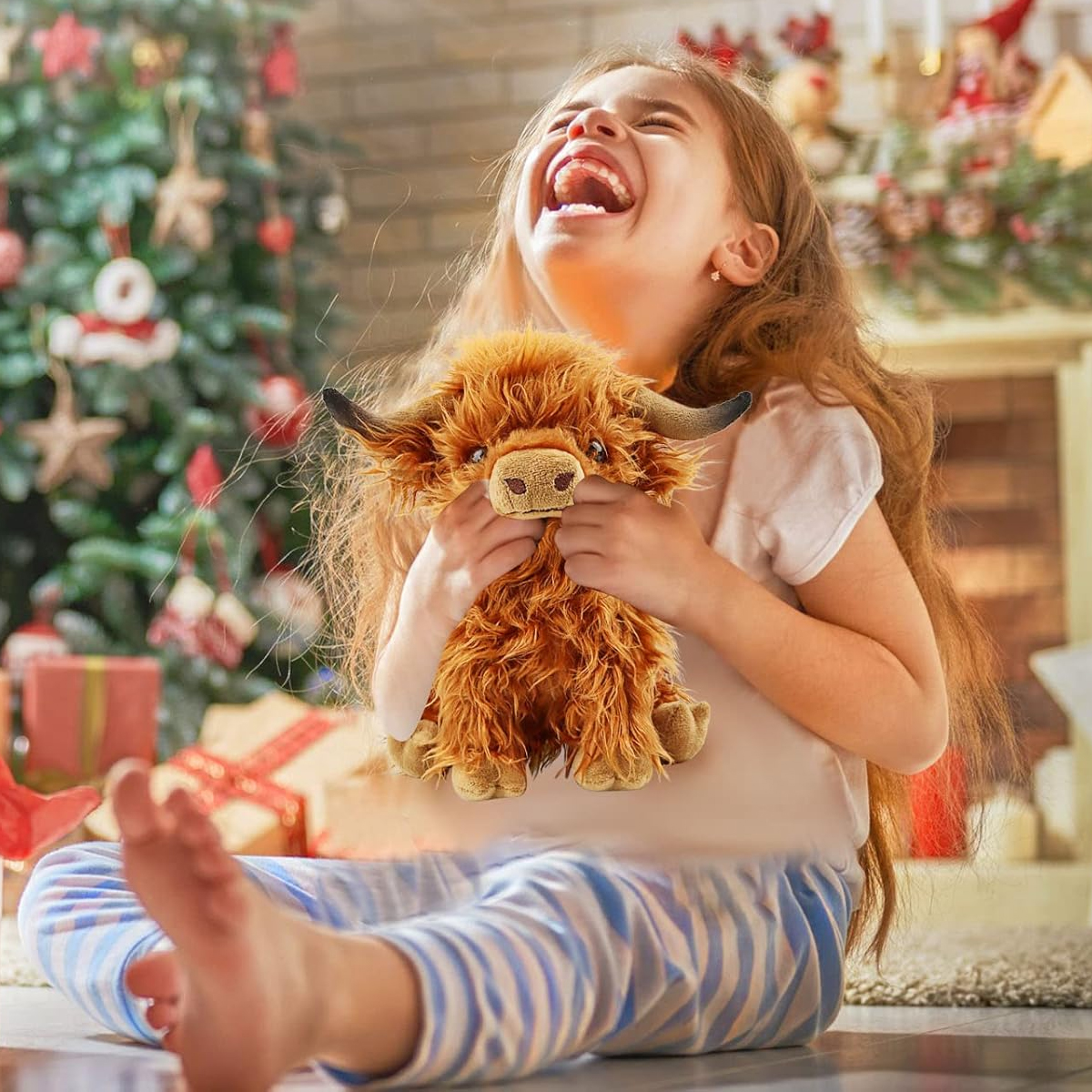 Ein Mädchen lacht, während es ein Stofftier, eine Hochlandkuh, hält. Im Hintergrund sind ein Weihnachtsbaum und ein Kamin zu sehen.