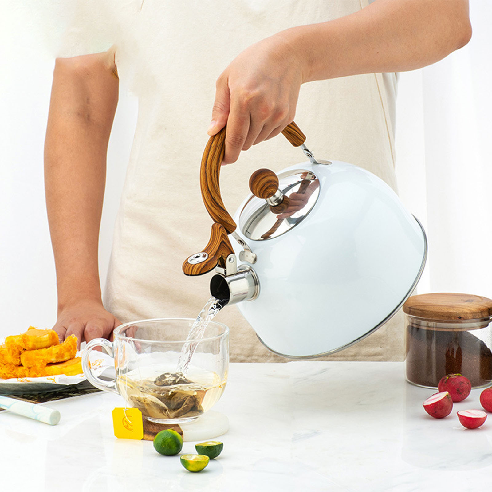 Eine Person gießt Wasser aus einem weißen Wasserkocher in ein Glas. Tee, Essen und Radieschen stehen auf dem Tisch.