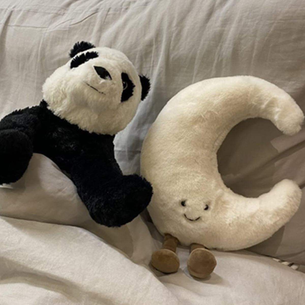Ein Panda-Plüschtier und ein Halbmond-Plüschtier liegen auf einem Bett. Der Panda ist schwarz und weiß, der Mond cremefarben mit kleinen Beinen.