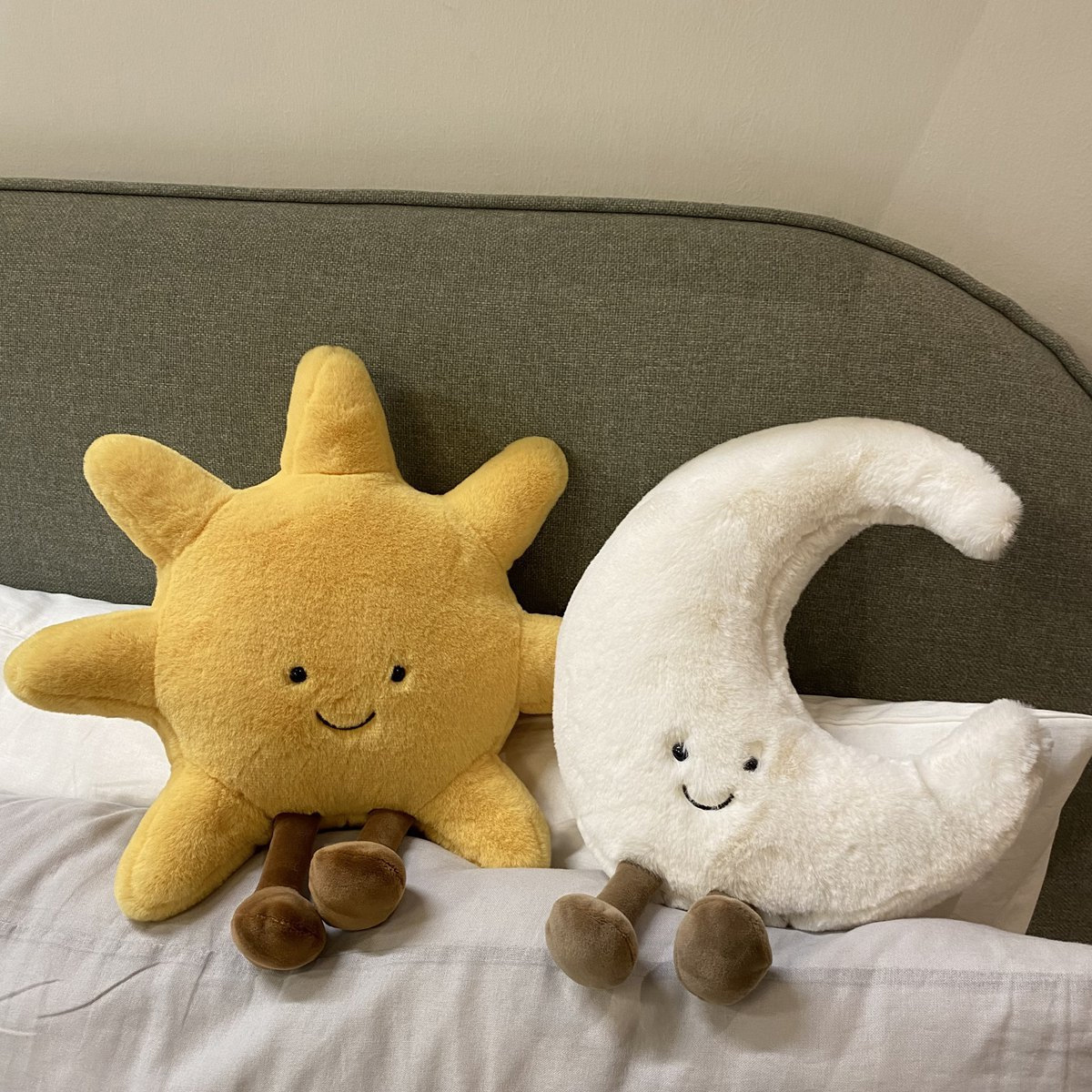 Ein Plüsch-Sonne und Mond mit lächelnden Gesichtern sitzen auf einem Bett. Die Sonne ist gelb, der Mond weiß. Dahinter ist ein graues Kopfteil.