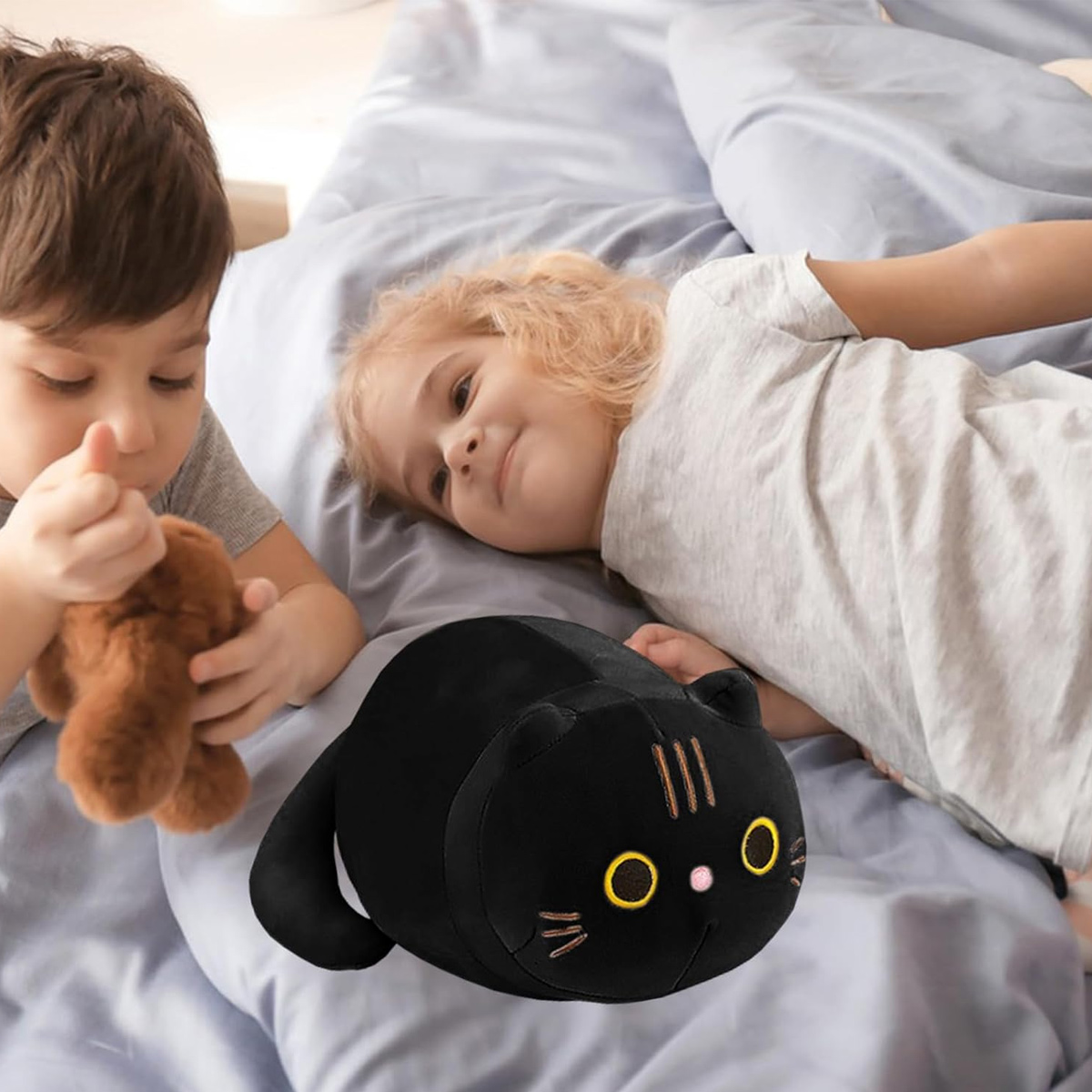 Zwei Kinder auf einem Bett mit einem schwarzen Katzenplüschtier. Ein Kind hält einen braunen Teddybären. Das andere Kind lächelt.