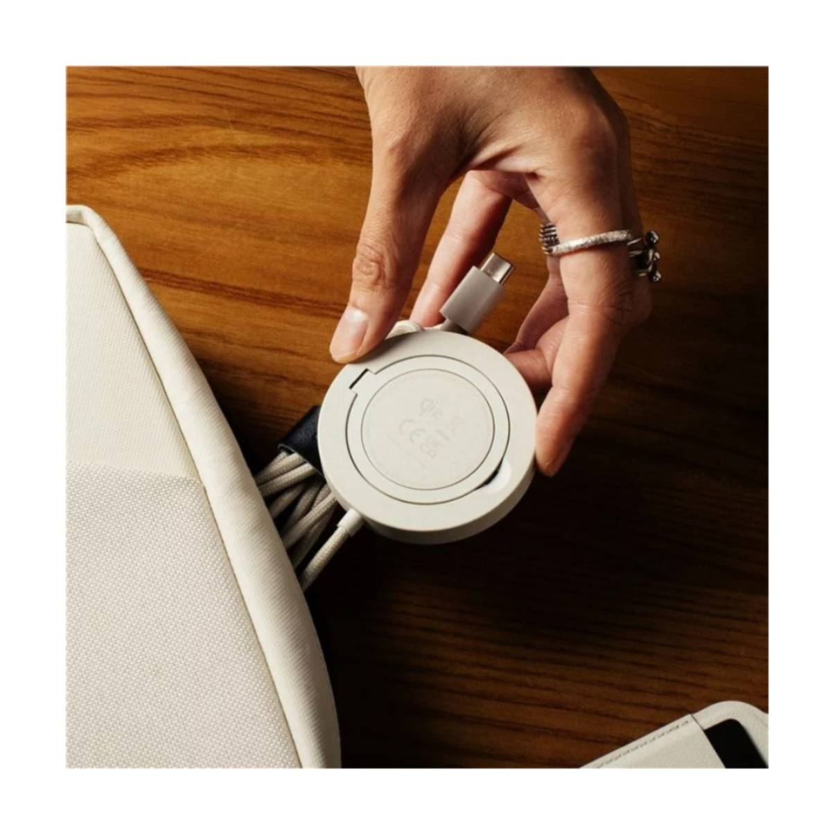 Eine Hand hält ein weißes, kreisförmiges Ladegerät in der Nähe einer cremefarbenen Tasche. Ringe zieren die Hand. Der Hintergrund ist eine Holzoberfläche.