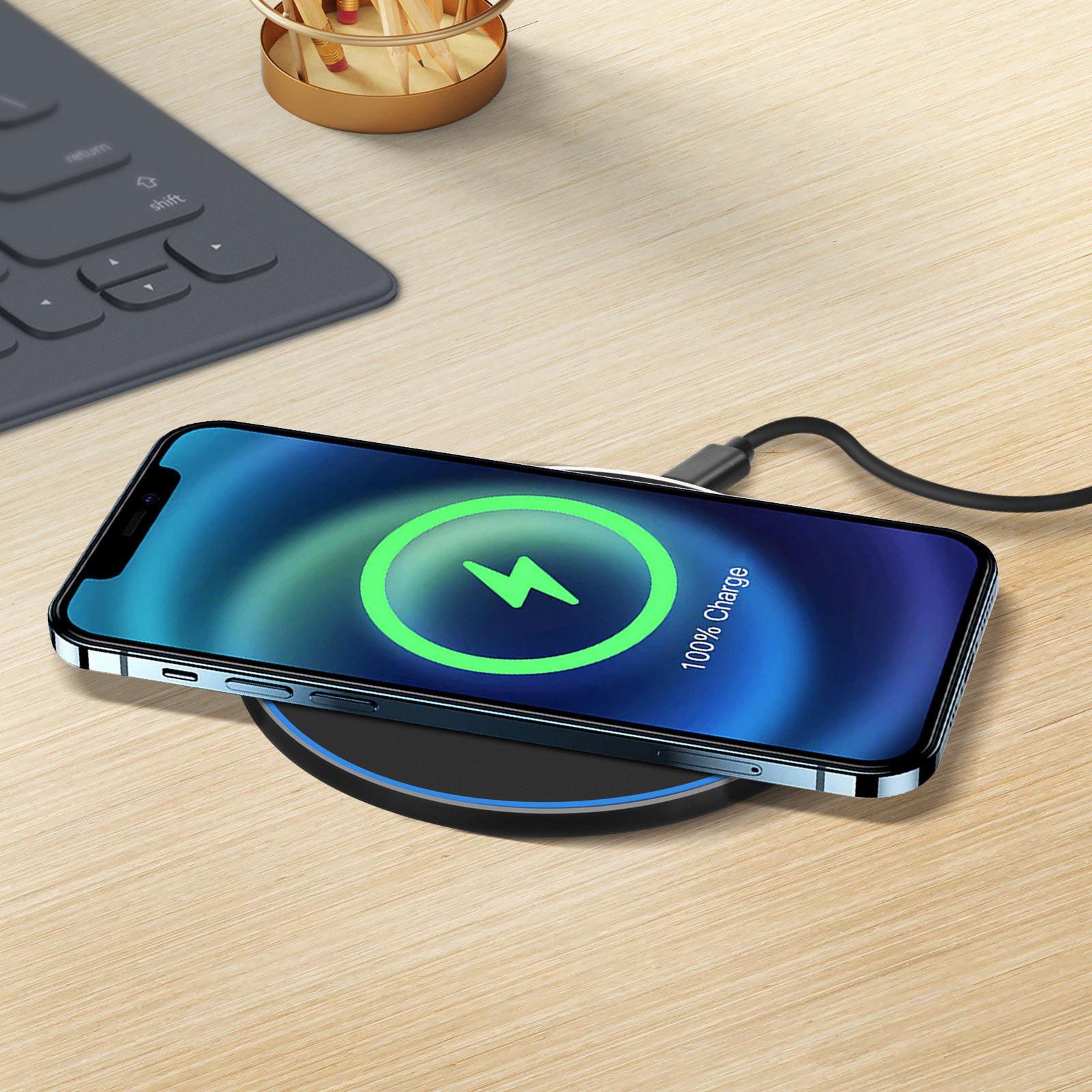 Un telefono si sta caricando in modalità wireless su un pad. Lo schermo mostra la carica al 100%. Tastiera e matite sullo sfondo.