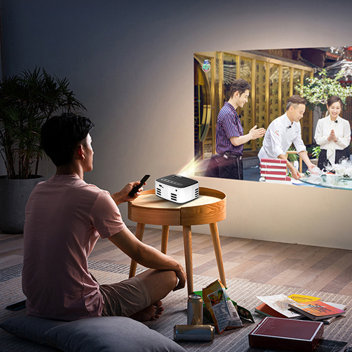 Ein Mann sitzt und schaut sich ein projiziertes Bild an einer Wand an. Ein Projektor steht auf einem Tisch. Das Bild zeigt Leute in einem Restaurant.