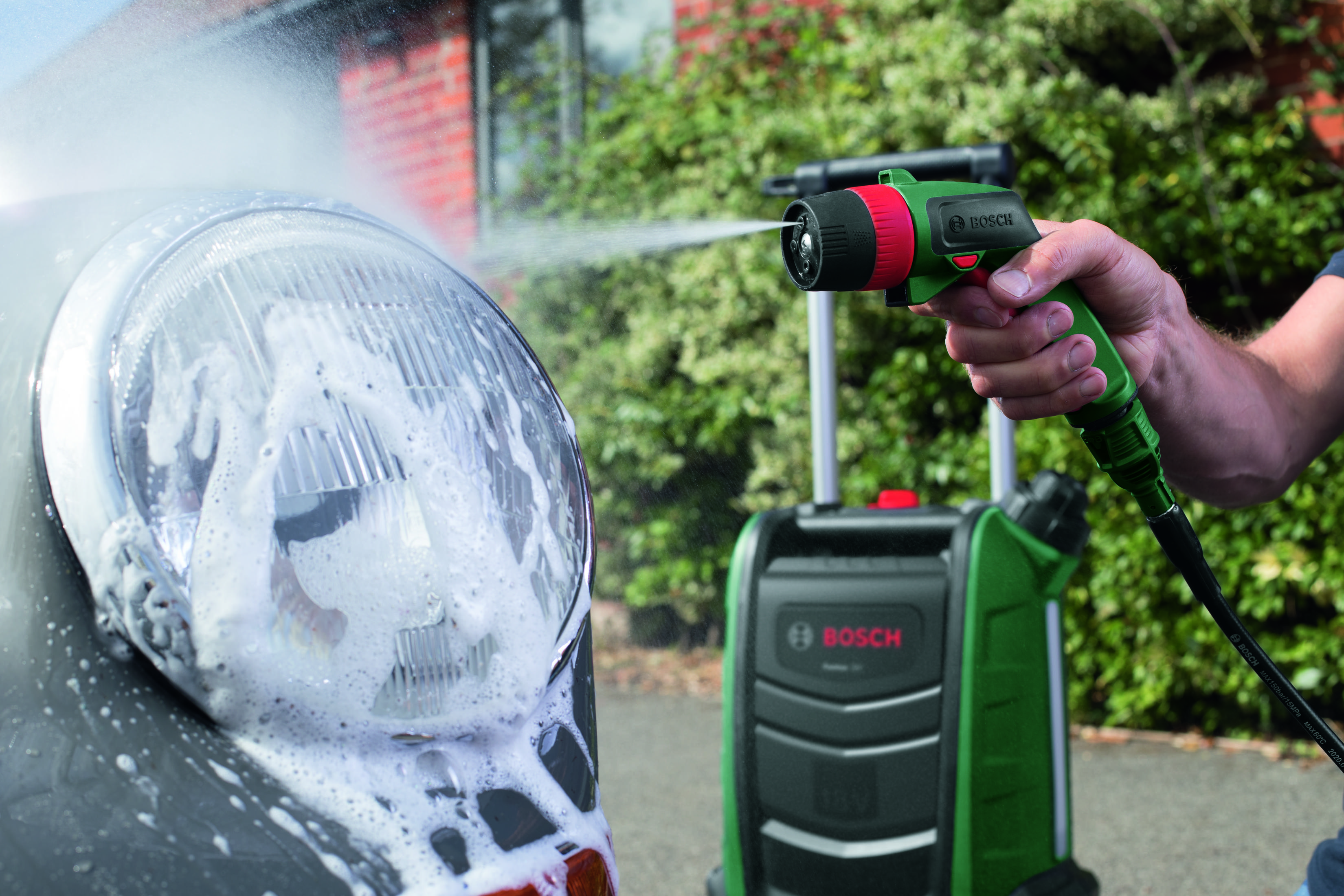 Une personne lave un phare de voiture avec un nettoyeur haute pression Bosch vert et rouge, pulvérisant de l'eau savonneuse.