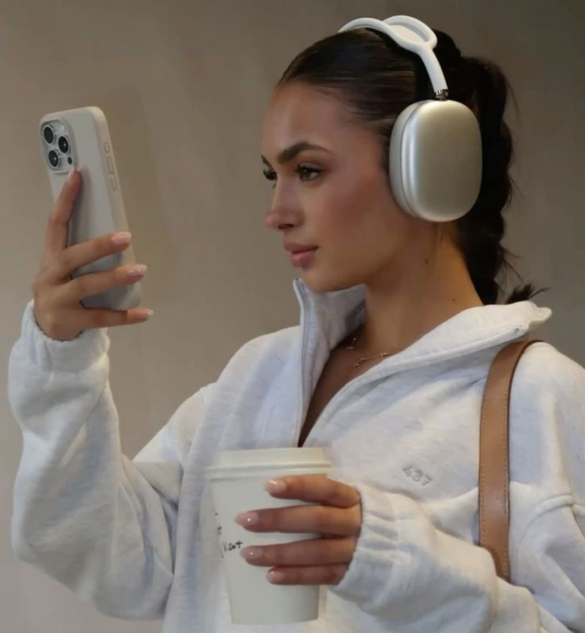 Mujer sosteniendo teléfono y café, usando auriculares y una sudadera blanca. El fondo es una pared neutral.