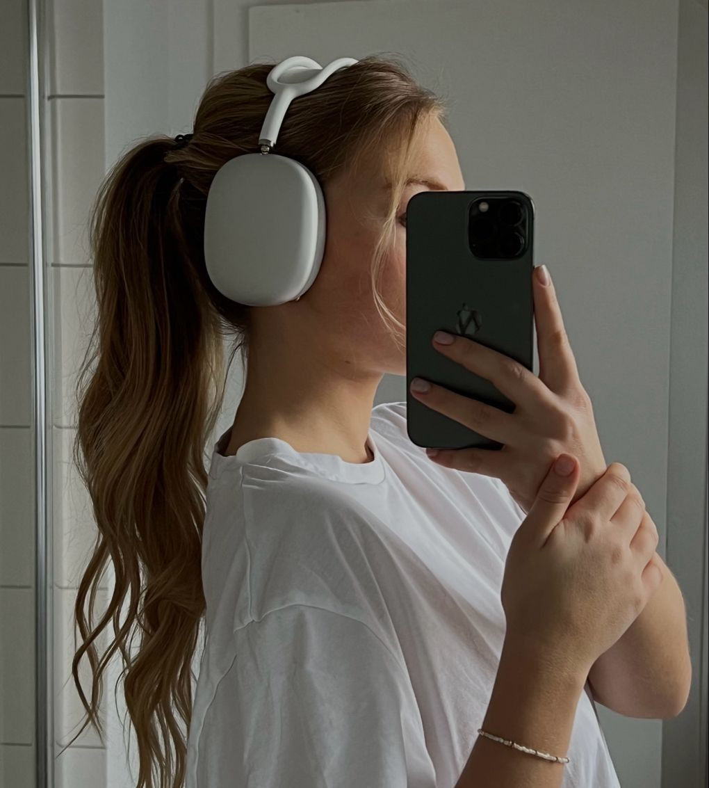 Una mujer con auriculares y un teléfono se hace un selfie en el espejo. Lleva una camisa blanca. Pelo largo, brazo con pulsera.
