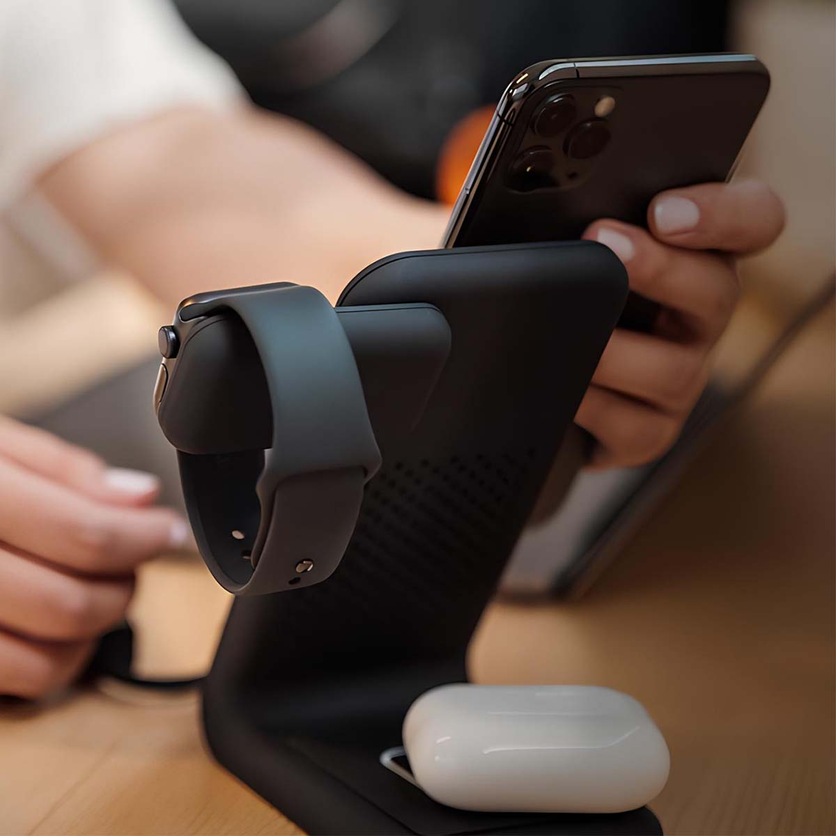 Eine Person hält ein Telefon auf einer schwarzen Ladestation. Eine Apple Watch und AirPods befinden sich ebenfalls auf dem Ladegerät.