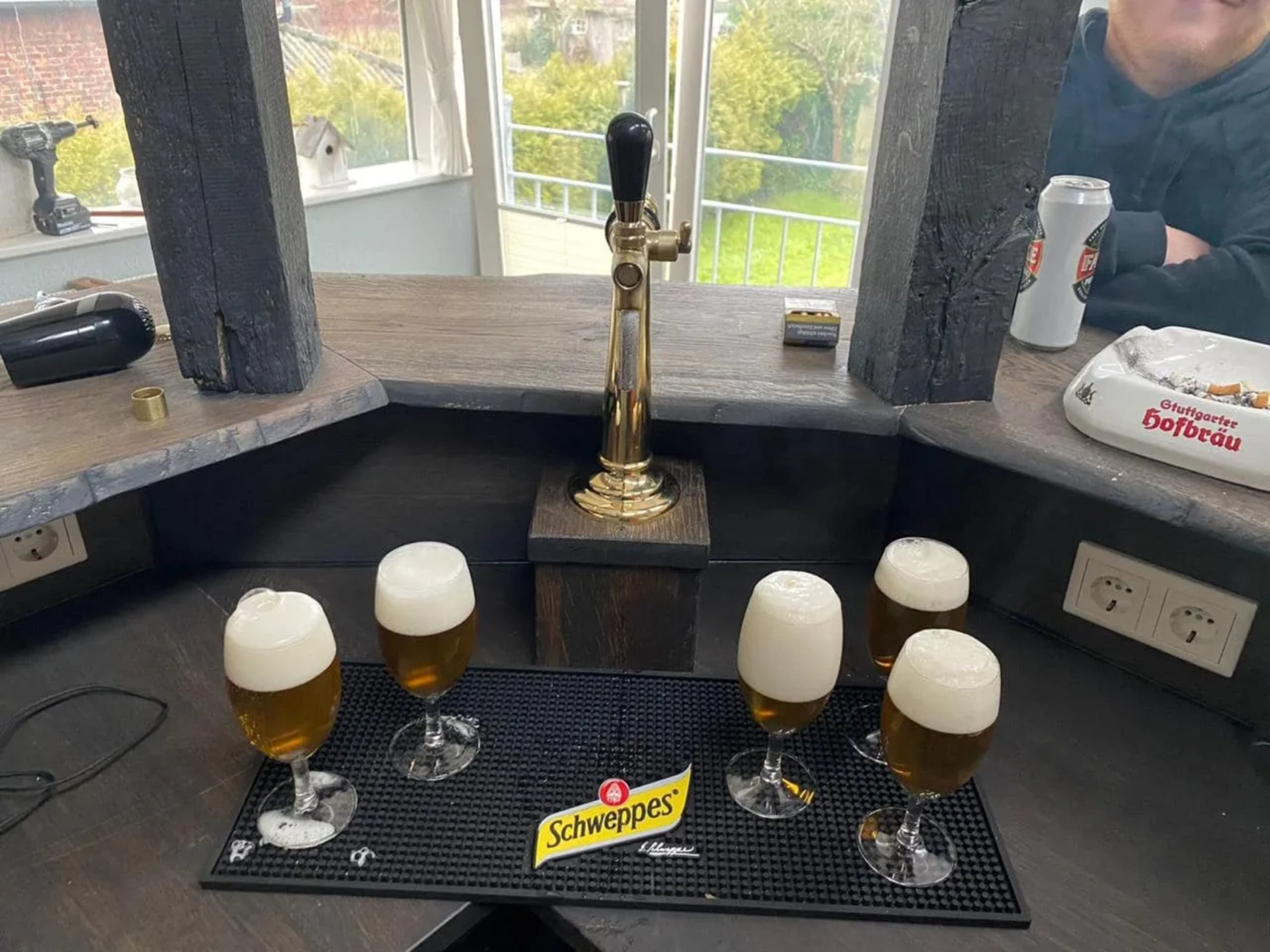 Eine Bartheke mit Biergläsern, einem Zapfhahn und anderen Gegenständen. Im Hintergrund sind ein Fenster und eine Person zu sehen.