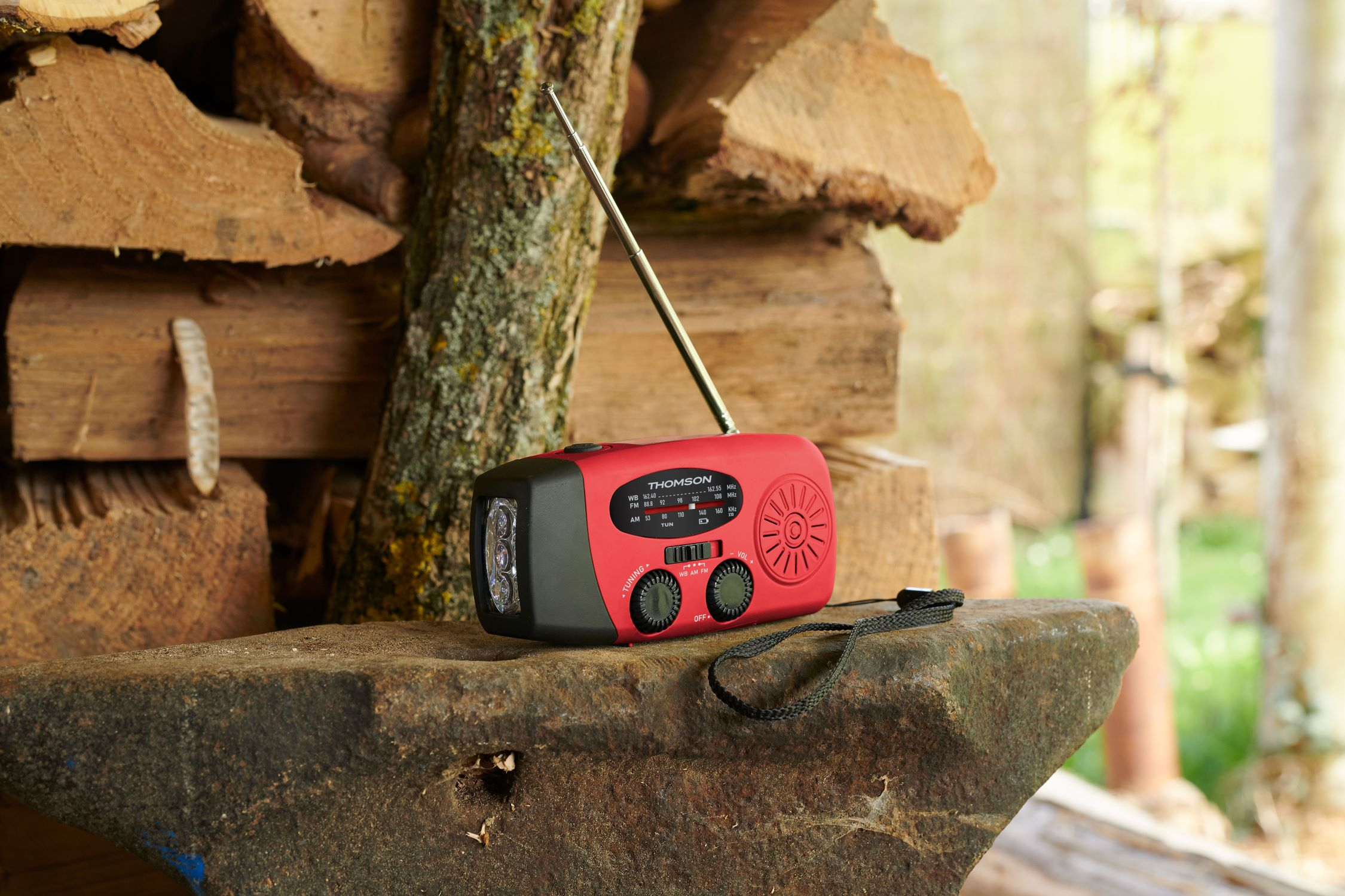 Una radio Thomson roja con frente negro, antena y correa está sobre una piedra. Madera en el fondo.