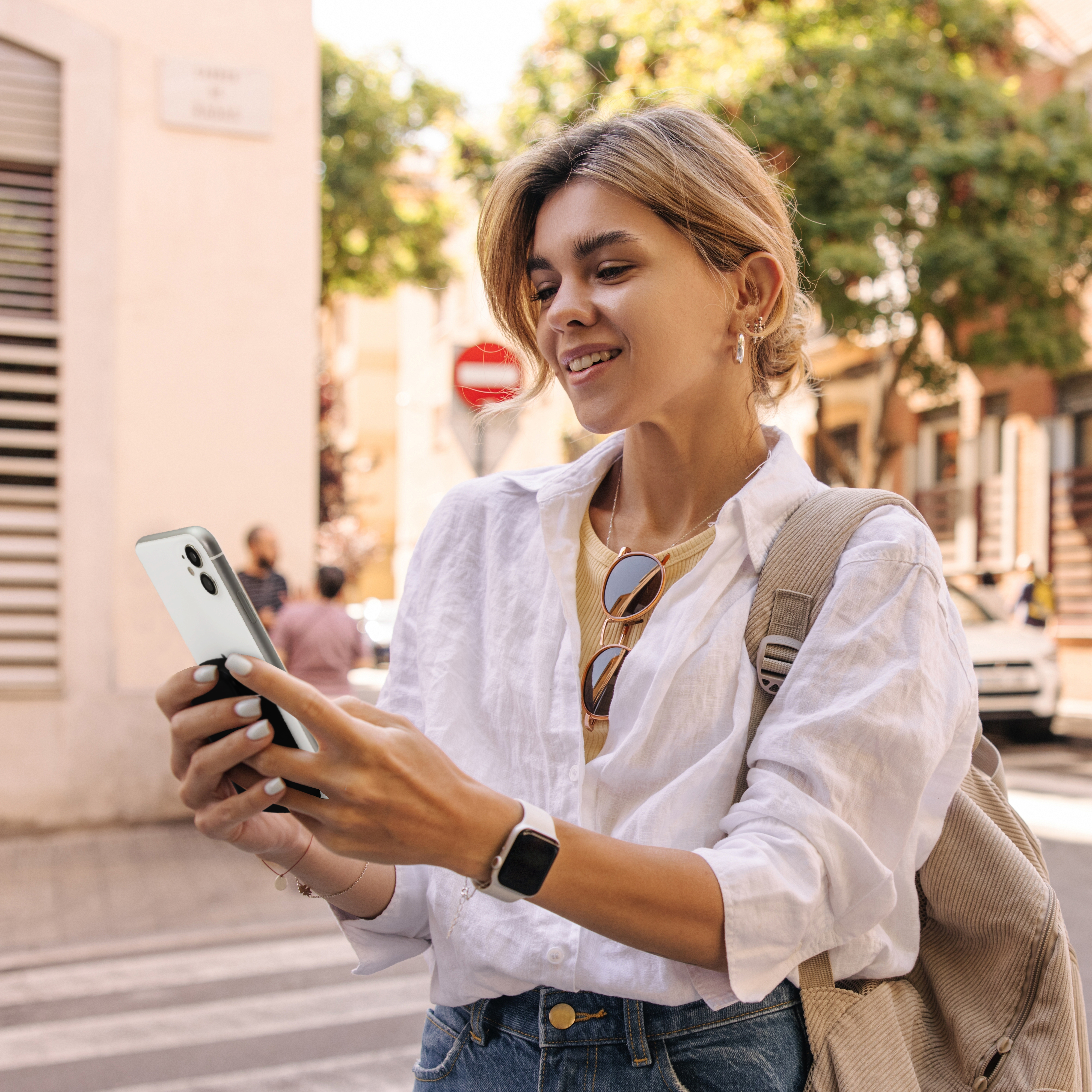 Una mujer mira su teléfono en la calle. Lleva camisa blanca, vaqueros y una mochila.