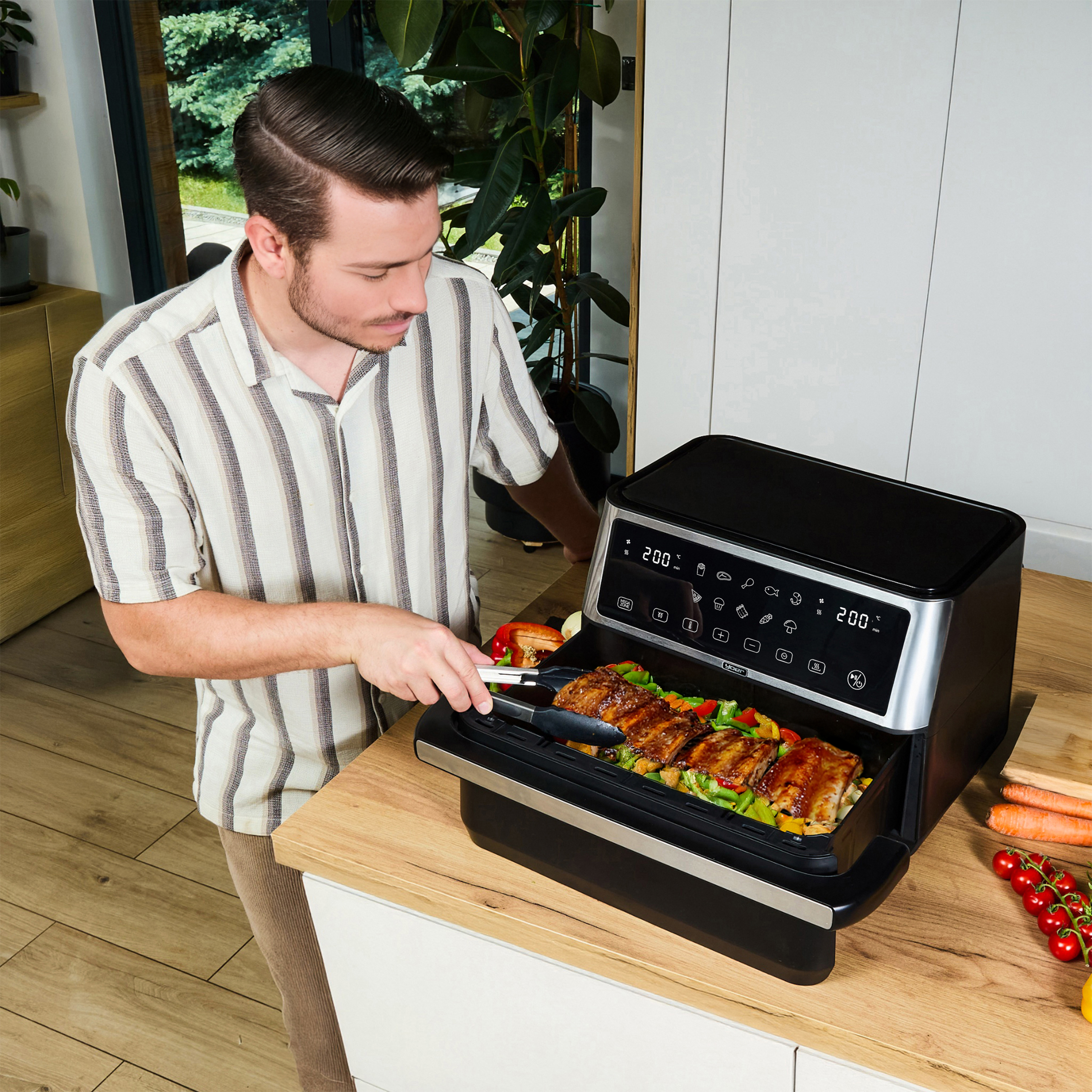 Mężczyzna używa szczypiec do wyjmowania jedzenia z frytkownicy powietrznej. Widoczne żeberka i warzywa. Kuchnia, drewniany blat.