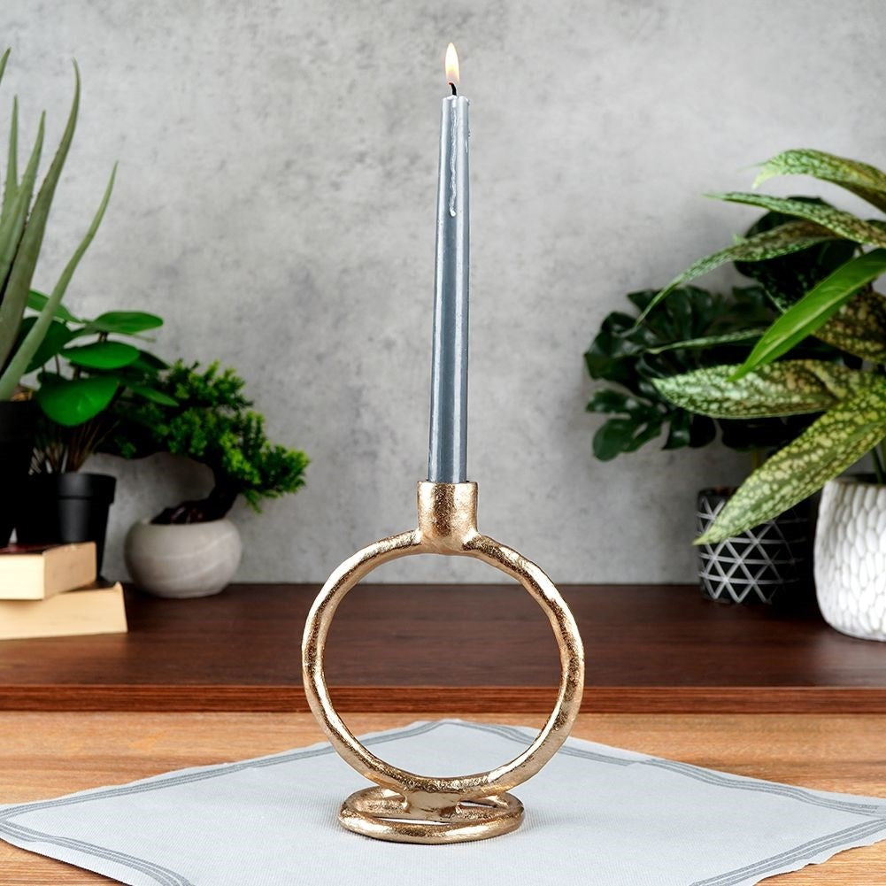 Una vela gris encendida en un soporte dorado en forma de anillo sobre un mantel gris claro. Plantas en macetas al fondo.
