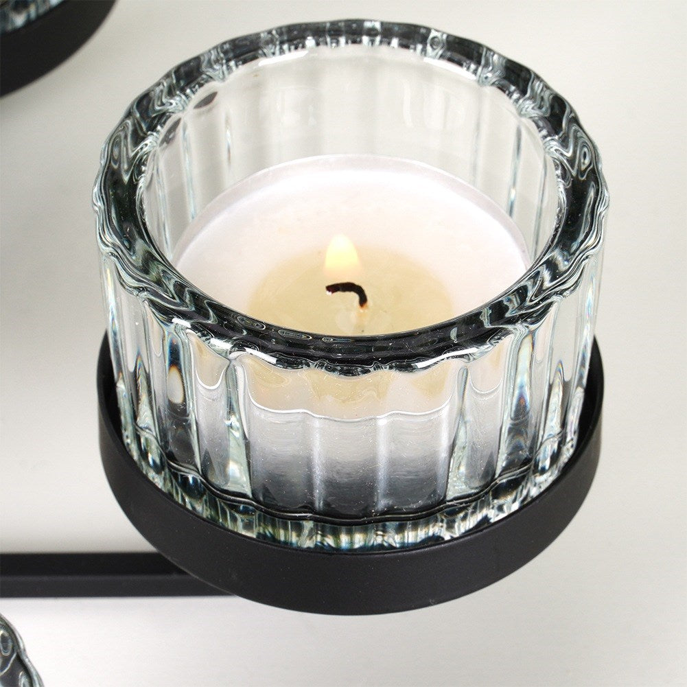 Una vela encendida en un portavelas de vidrio transparente sobre una base de metal negro. La llama es amarilla y el fondo blanco.