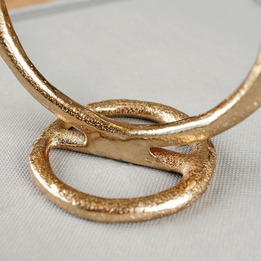 Primer plano de un anillo de metal dorado y texturizado entrelazado con otro anillo, sobre un fondo de tela gris claro.