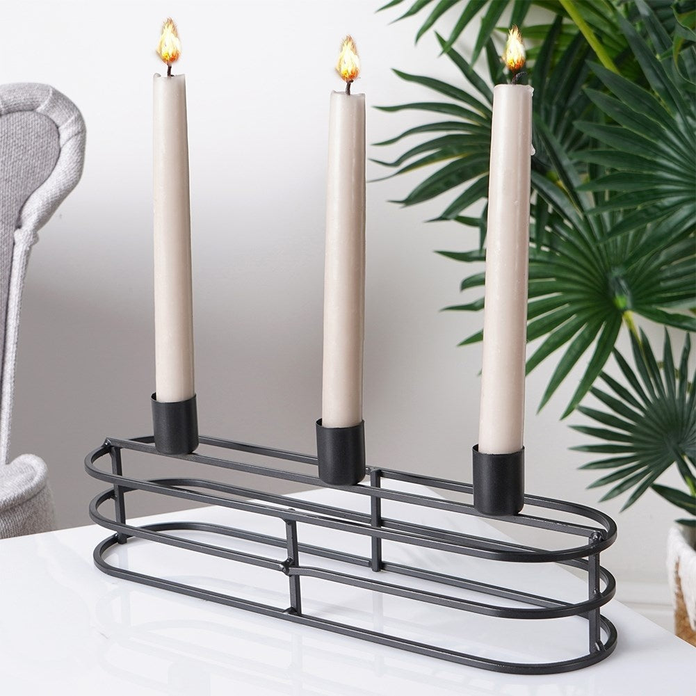 Tres velas encendidas en un soporte de metal negro sobre una superficie blanca. Planta verde al fondo.