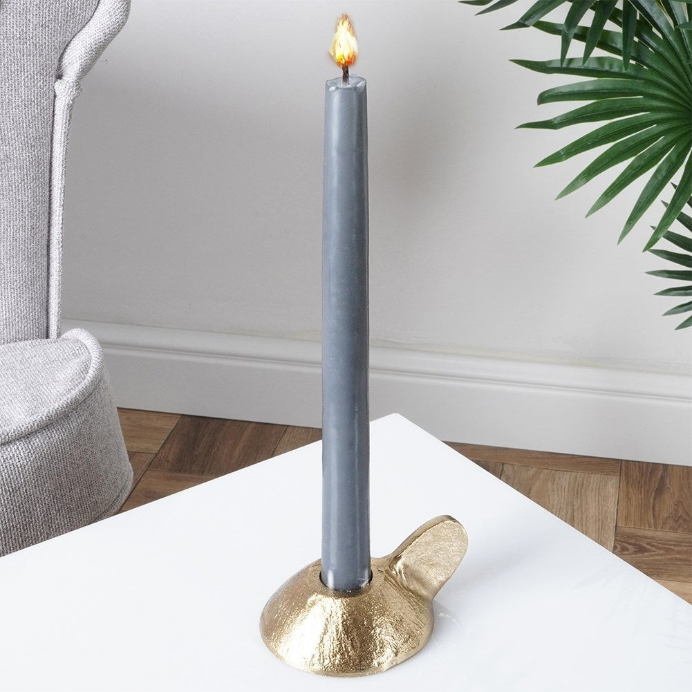 Una vela gris encendida en un soporte dorado sobre una mesa blanca. Una silla gris y una planta verde en el fondo.